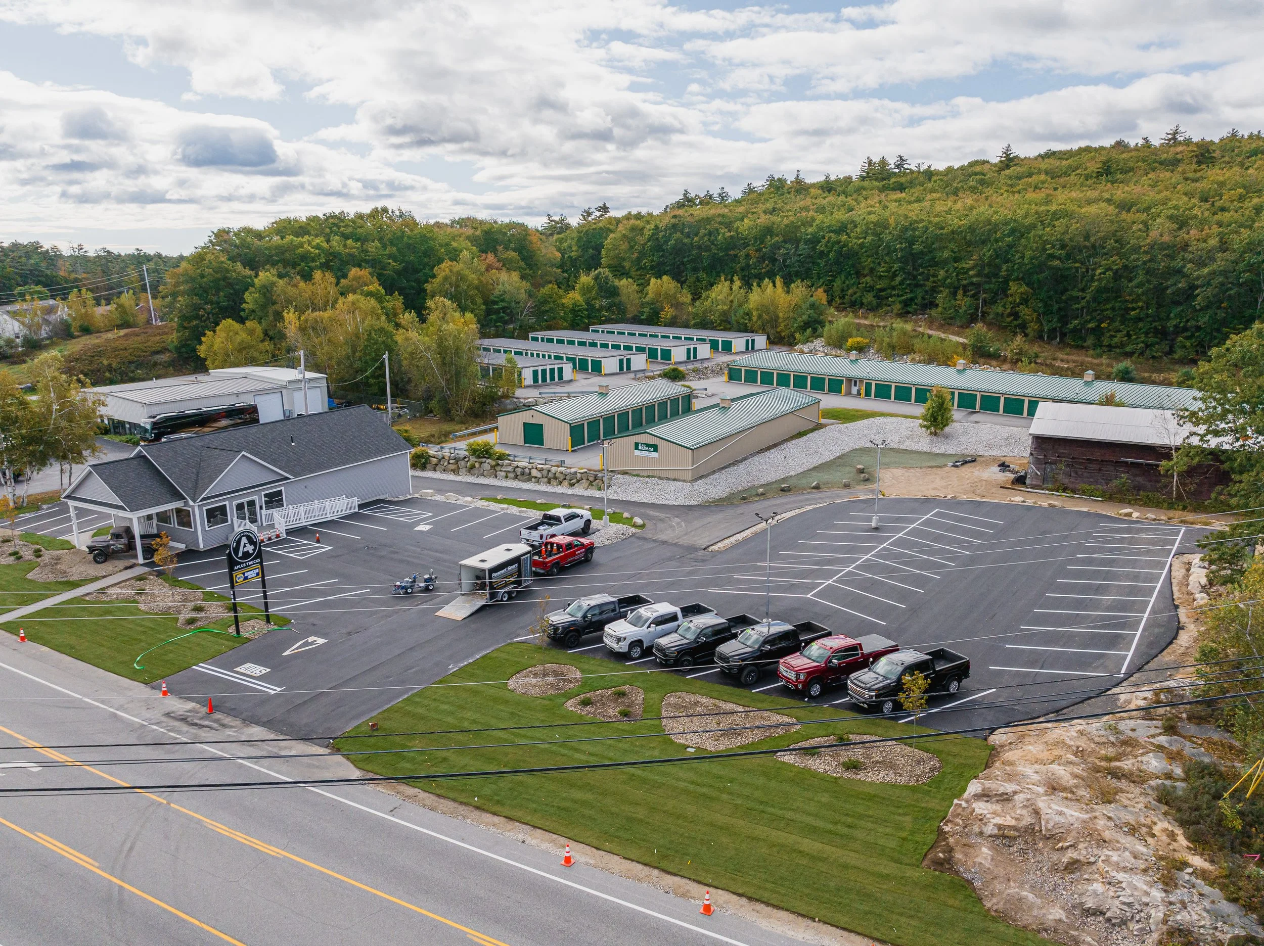 Aerial view of a small commercial area with a parking lot, some cars, a building with a sign, and storage facilities in a hilly and wooded landscape.