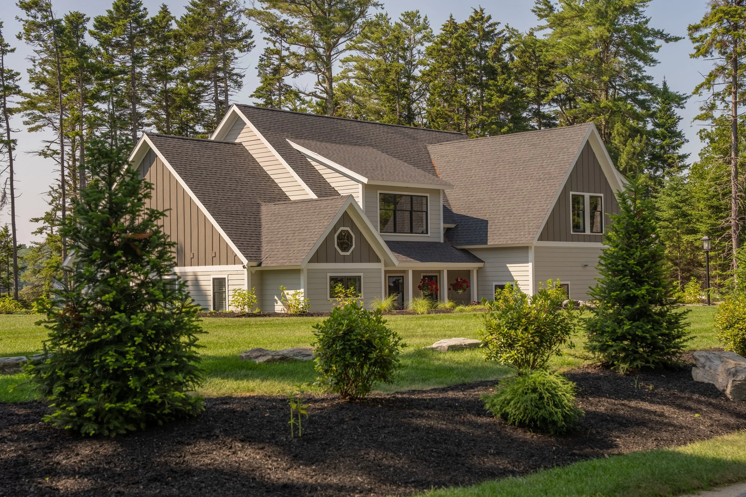 A modern house with multiple gabled roofs, white and dark gray siding, surrounded by a well-manicured lawn and small evergreen trees, with a wooded area in the background.