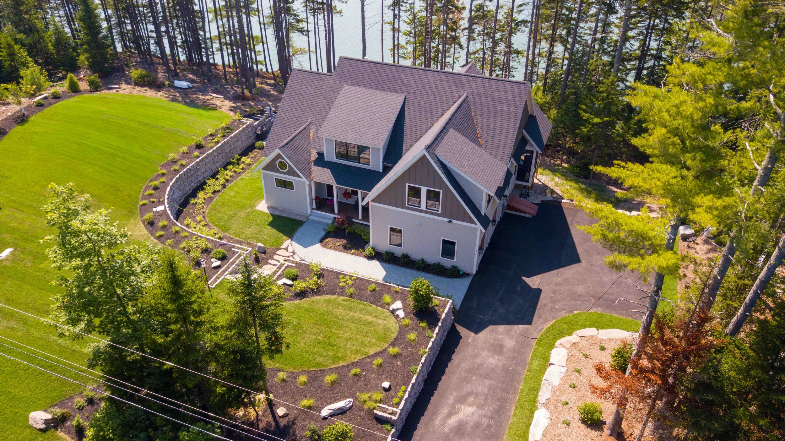 Aerial view of a modern house with a gabled roof, surrounded by a landscaped yard with grass, bushes, and trees, near a body of water.
