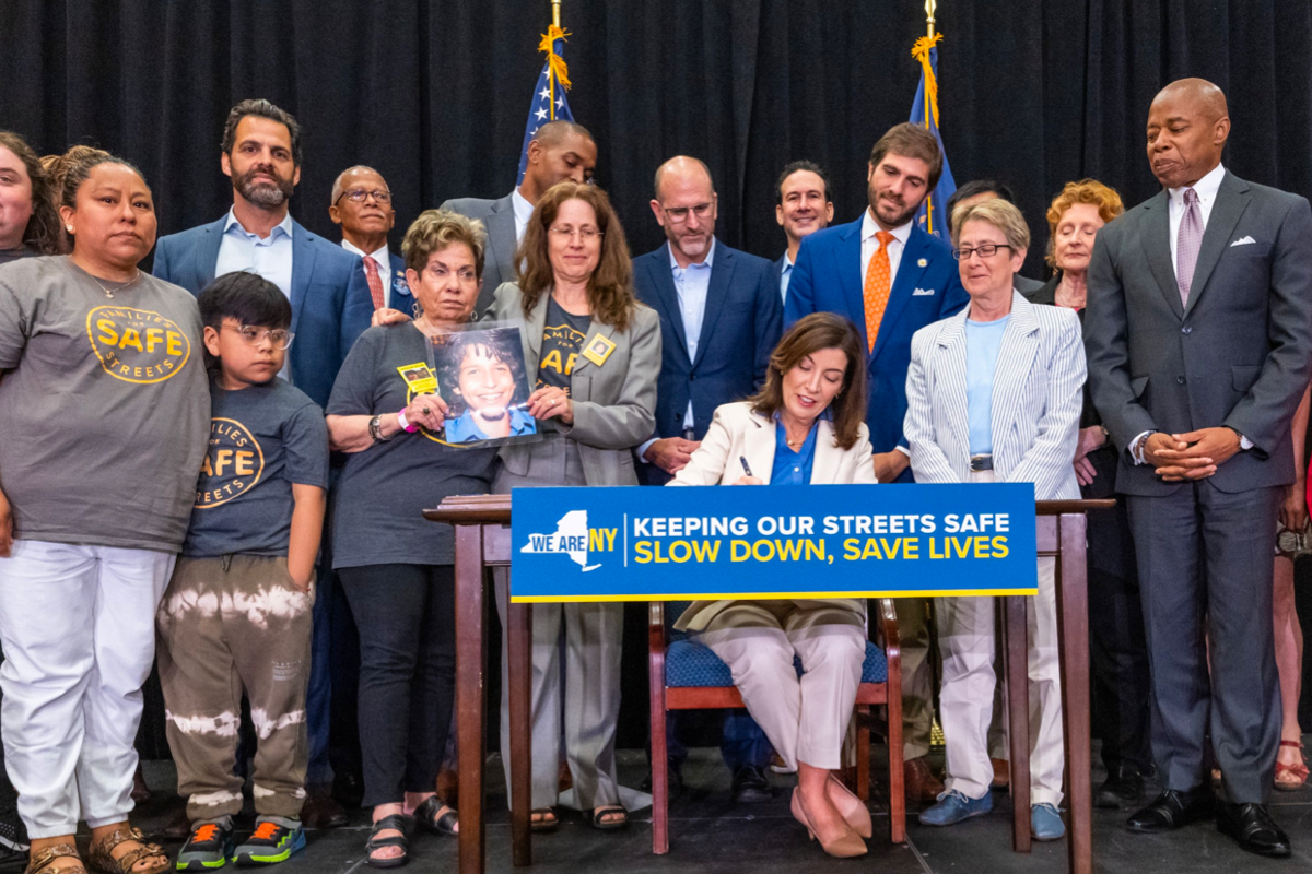Governor Kathy Hochul sitting at a table and signing a bill for expanded speed safety camera operation, surrounded by a crowd of safe streets advocates