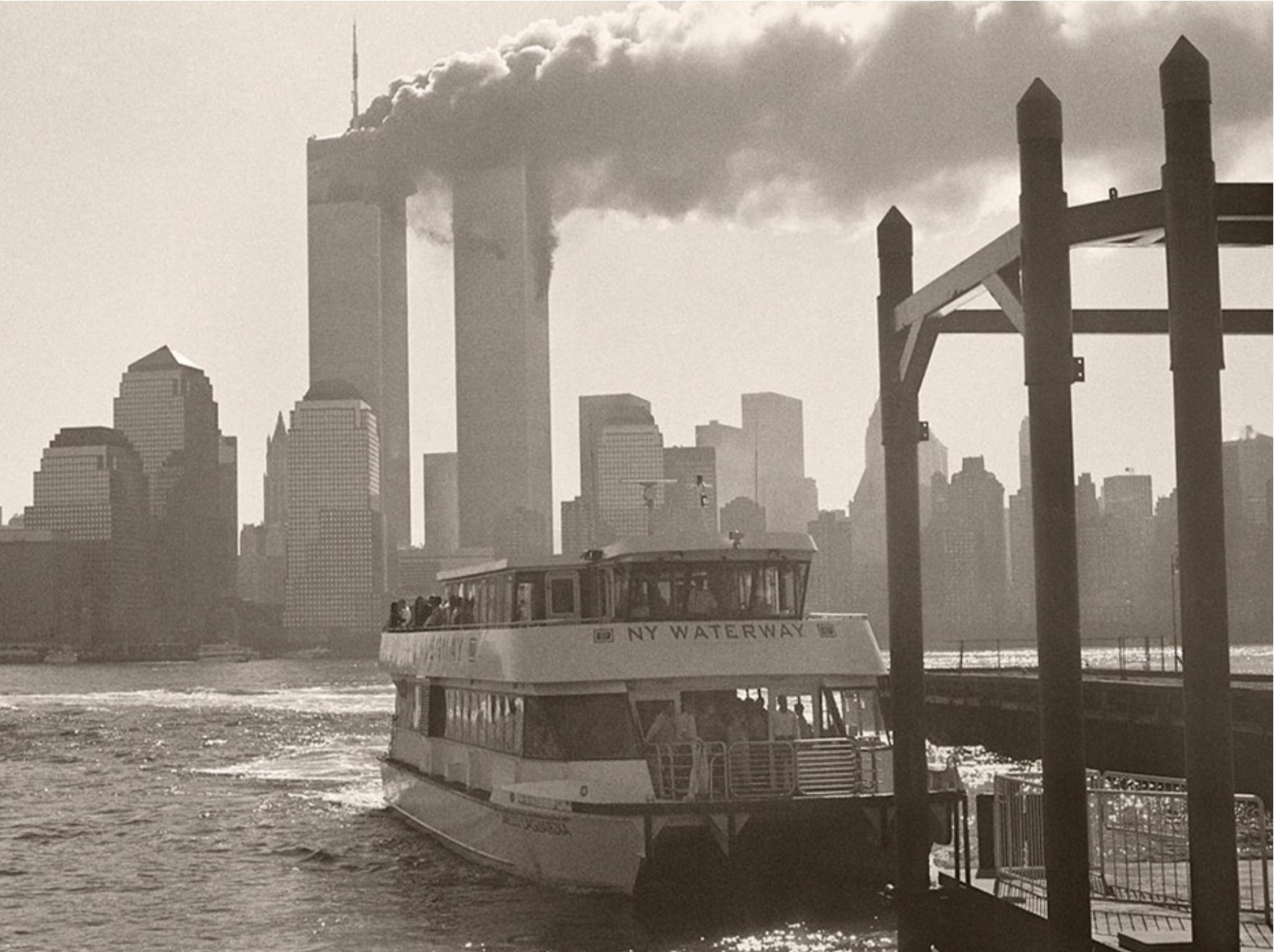A city skyline with tall buildings and smoke rising from two of the buildings, seen from the water. In the foreground, a boat labeled 'NY Waterway' is on the water, with a group of people on it.