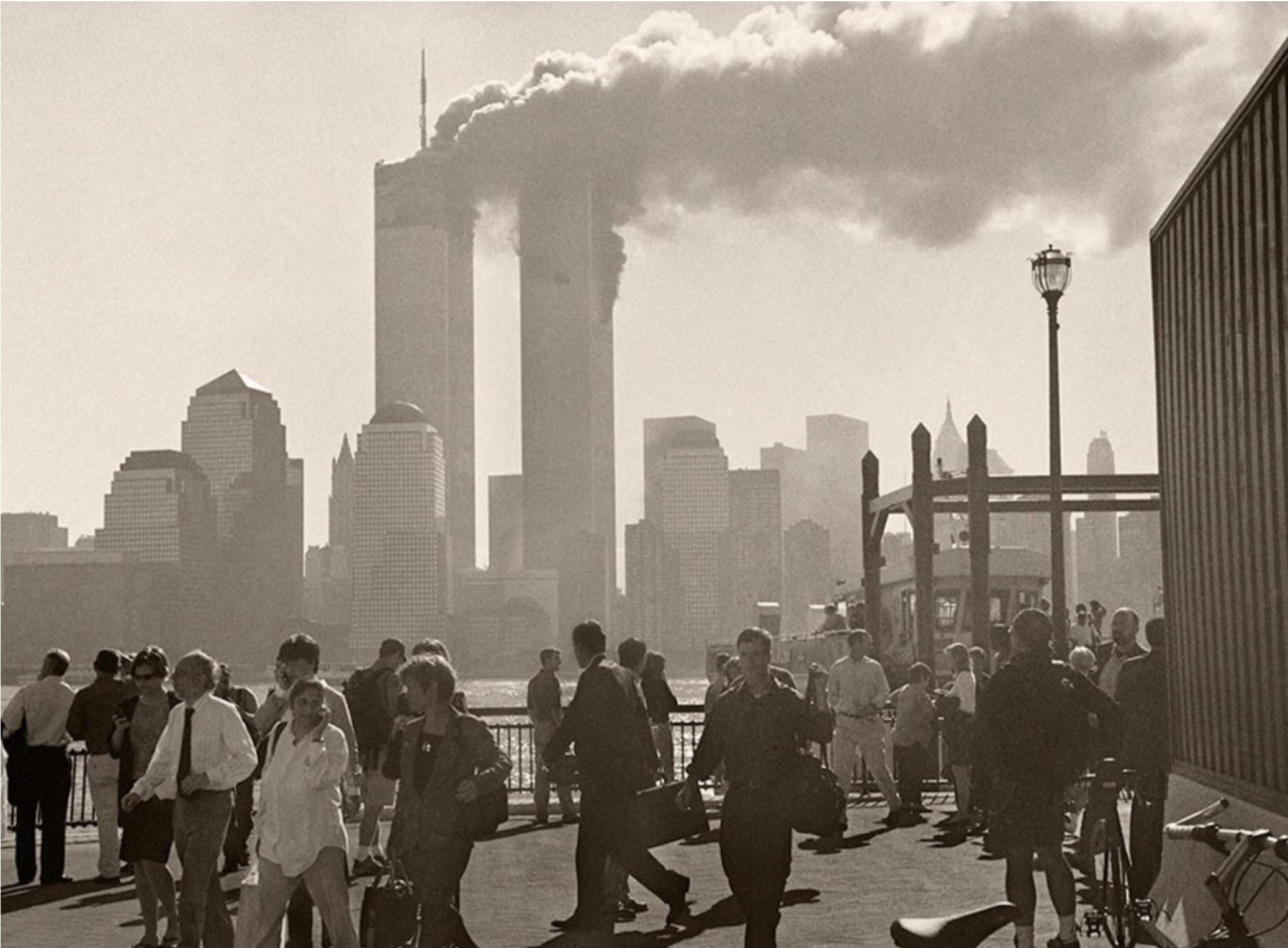 People standing and walking at a waterfront with the skyline of New York City, including the Twin Towers, in the background. Smoke billows from the Twin Towers.