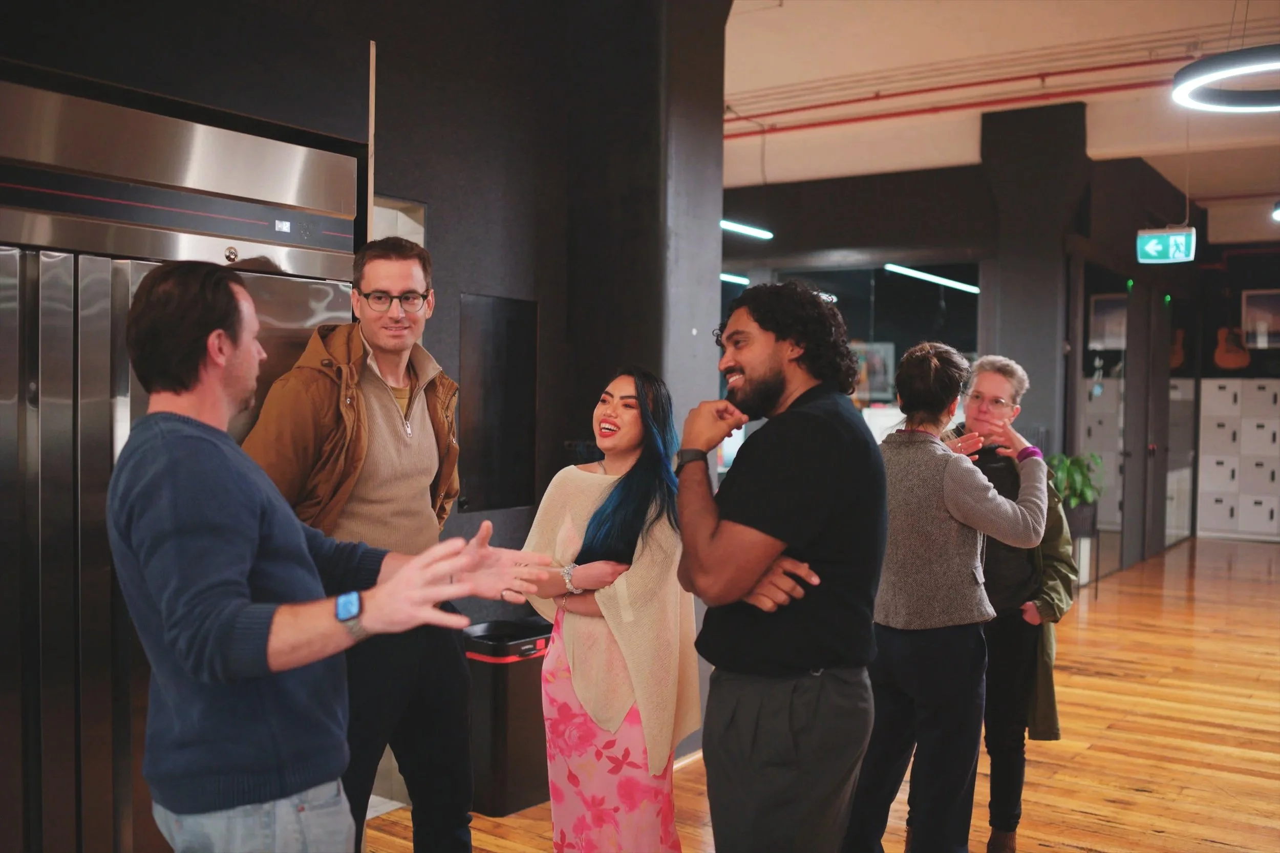 A group of five people talking and smiling in a modern indoor space, with some wearing glasses and casual clothing, and two women and three men visible.