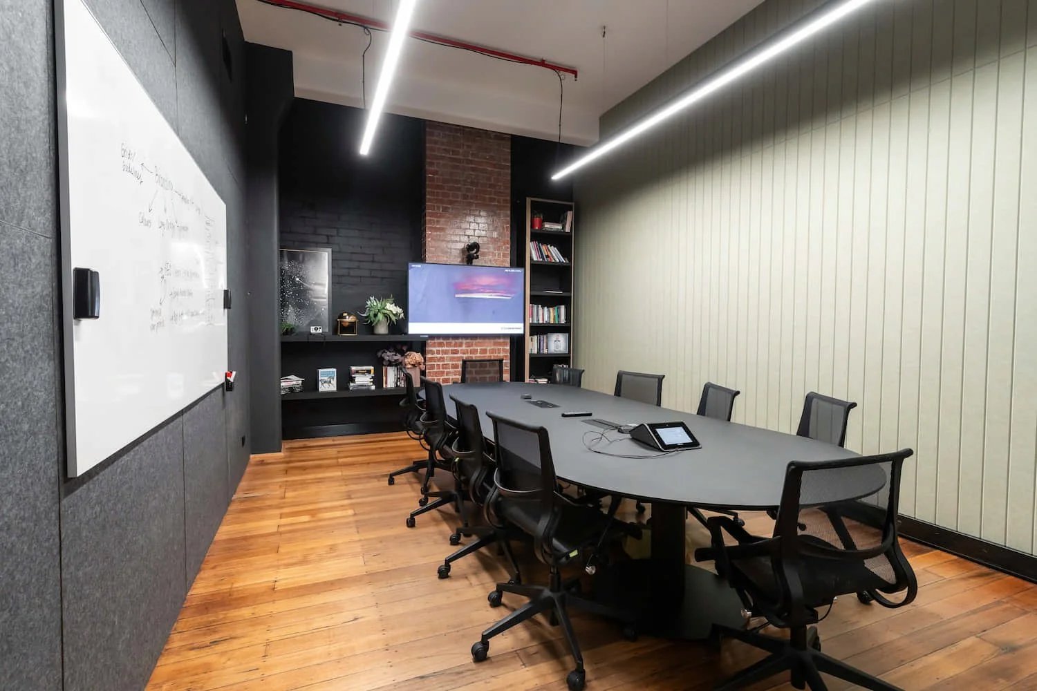 Modern conference room with a long black table, black office chairs, a whiteboard on one wall, bookshelves, a large screen, and decorative plants.