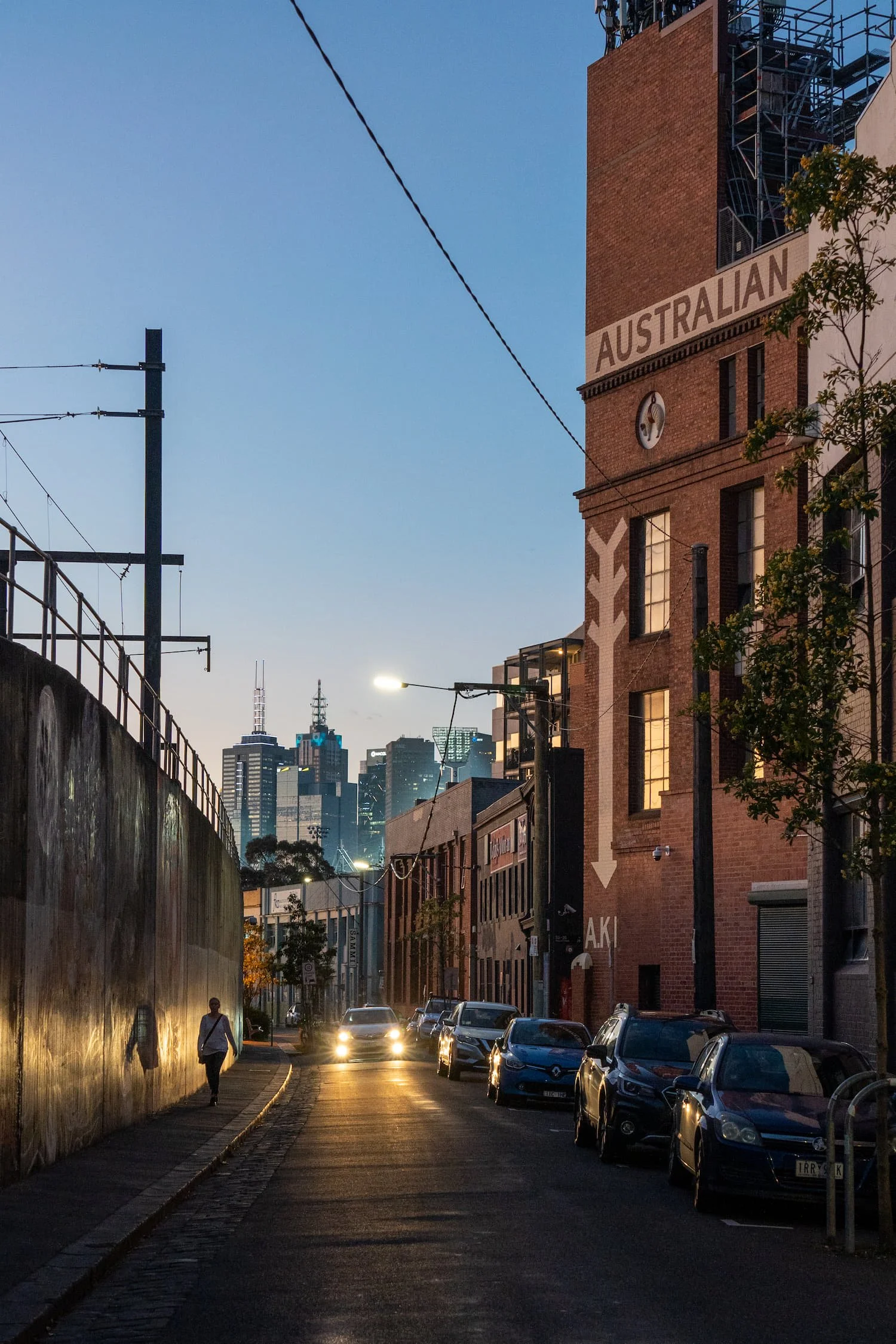 City street at dusk with parked cars along the right side, a brick building with a sign that says 'AUSTRALIAN,' and a skyline with tall buildings in the background. A person walks on the sidewalk on the left, and streetlights cast a warm glow.