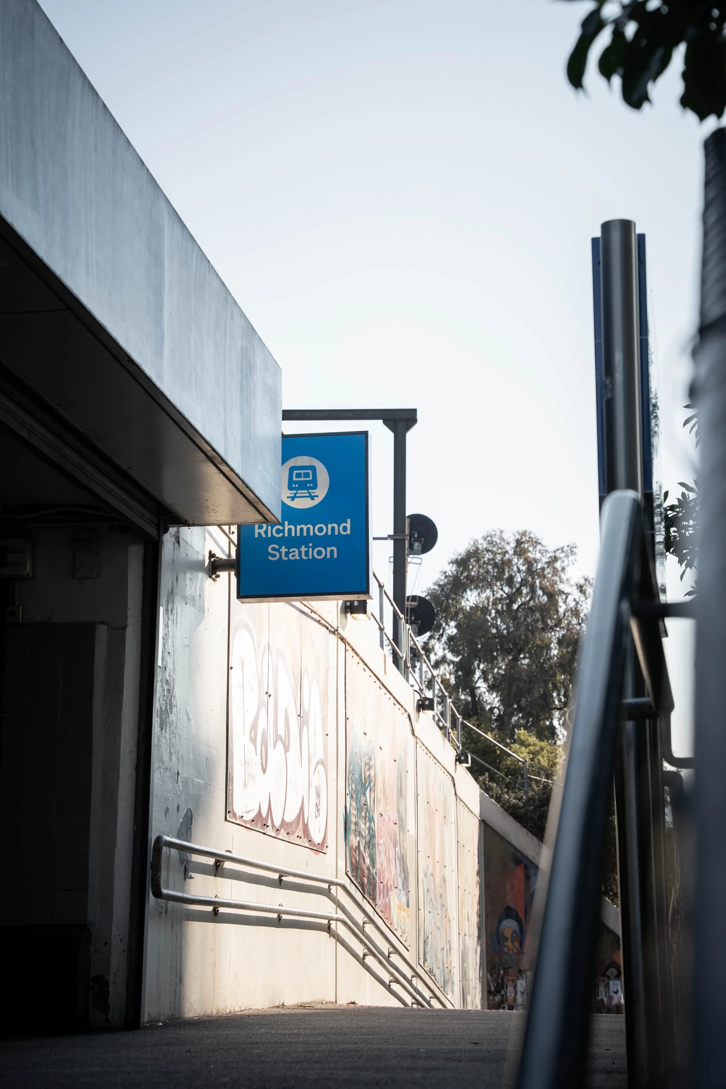 Street scene showing a sidewalk leading under an overpass with a Richmond Station sign, graffitied wall, and trees in the background, taken from a low angle.