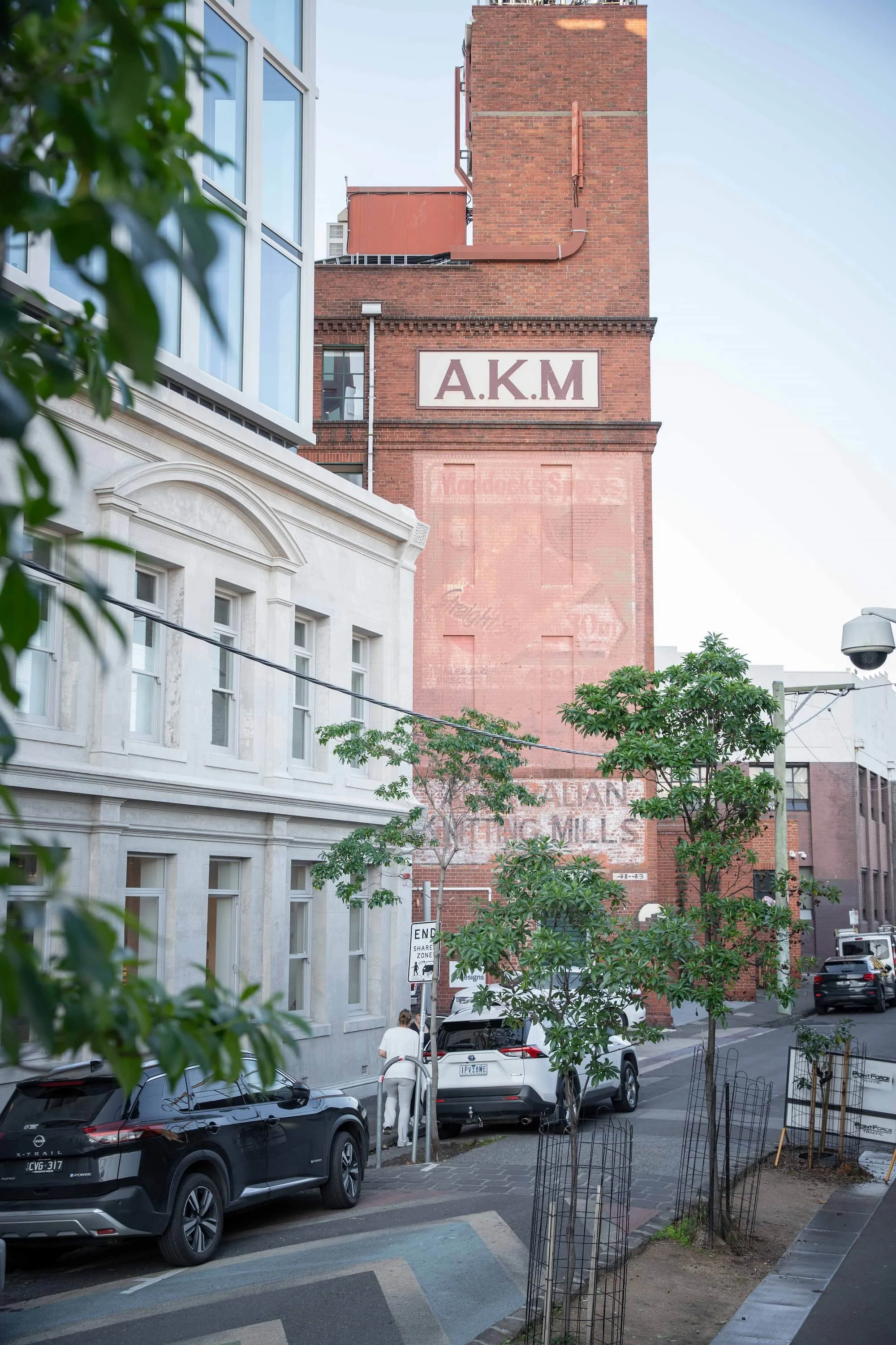Street scene showing parked cars, trees, a white building with windows, and a tall brick building with a sign reading 'A.K.M.'.