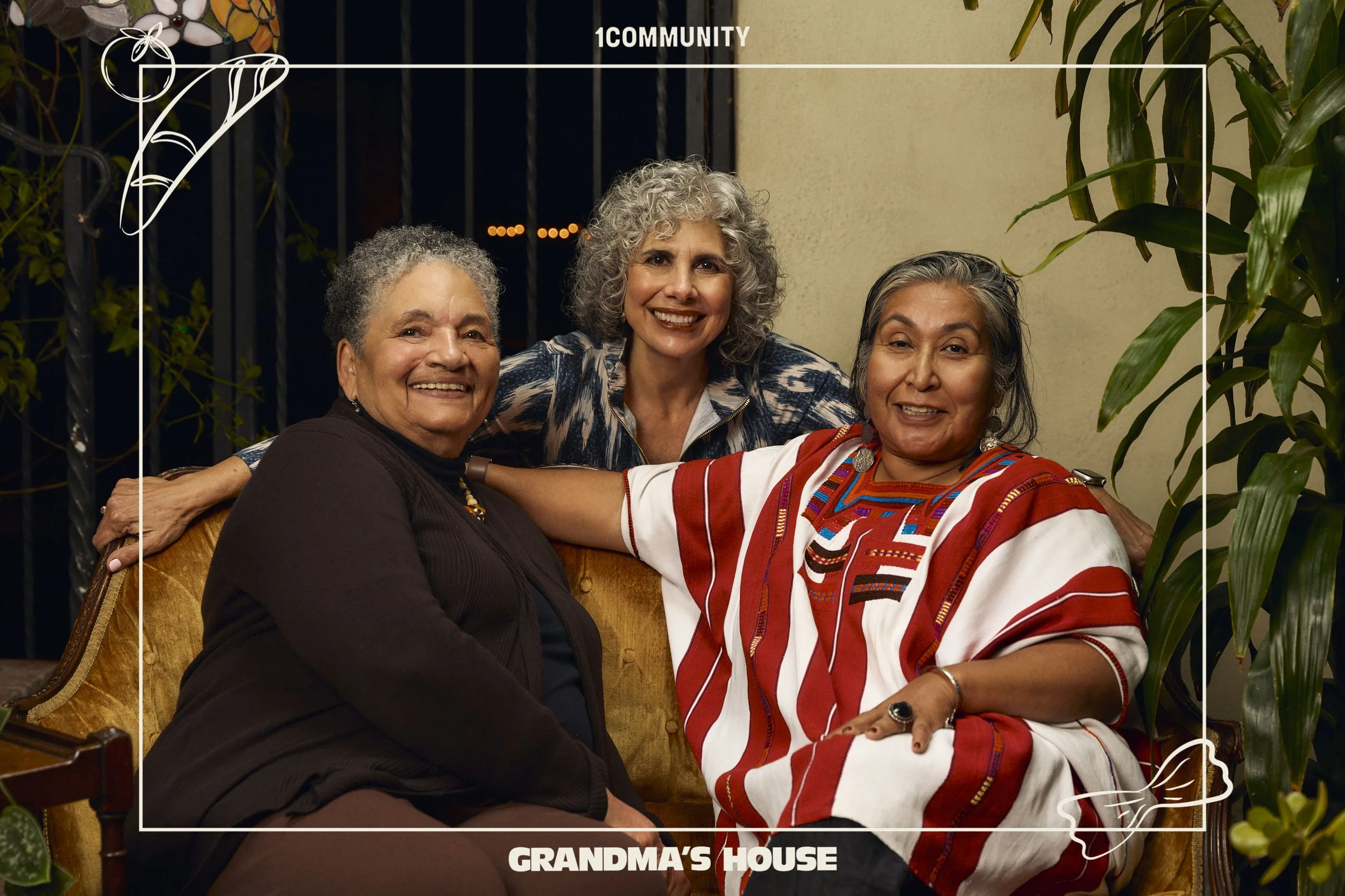 Three women sitting closely together on a vintage couch, smiling at the camera, in a cozy indoor setting with plants around and a metal gate in the background. The text overlays say '1Community' at the top and 'Grandma's House' at the bottom.