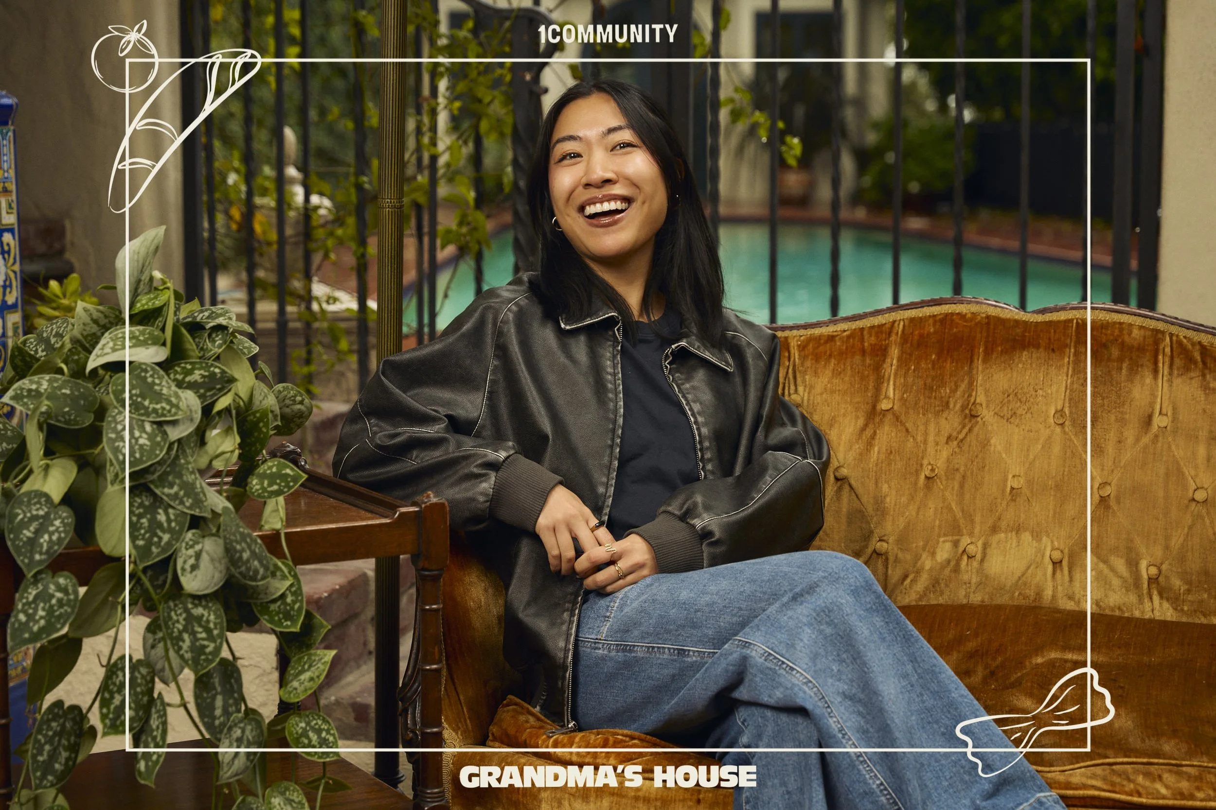A woman sitting on a vintage sofa outside near a swimming pool, smiling, wearing a black leather jacket and blue jeans, surrounded by greenery and potted plants.