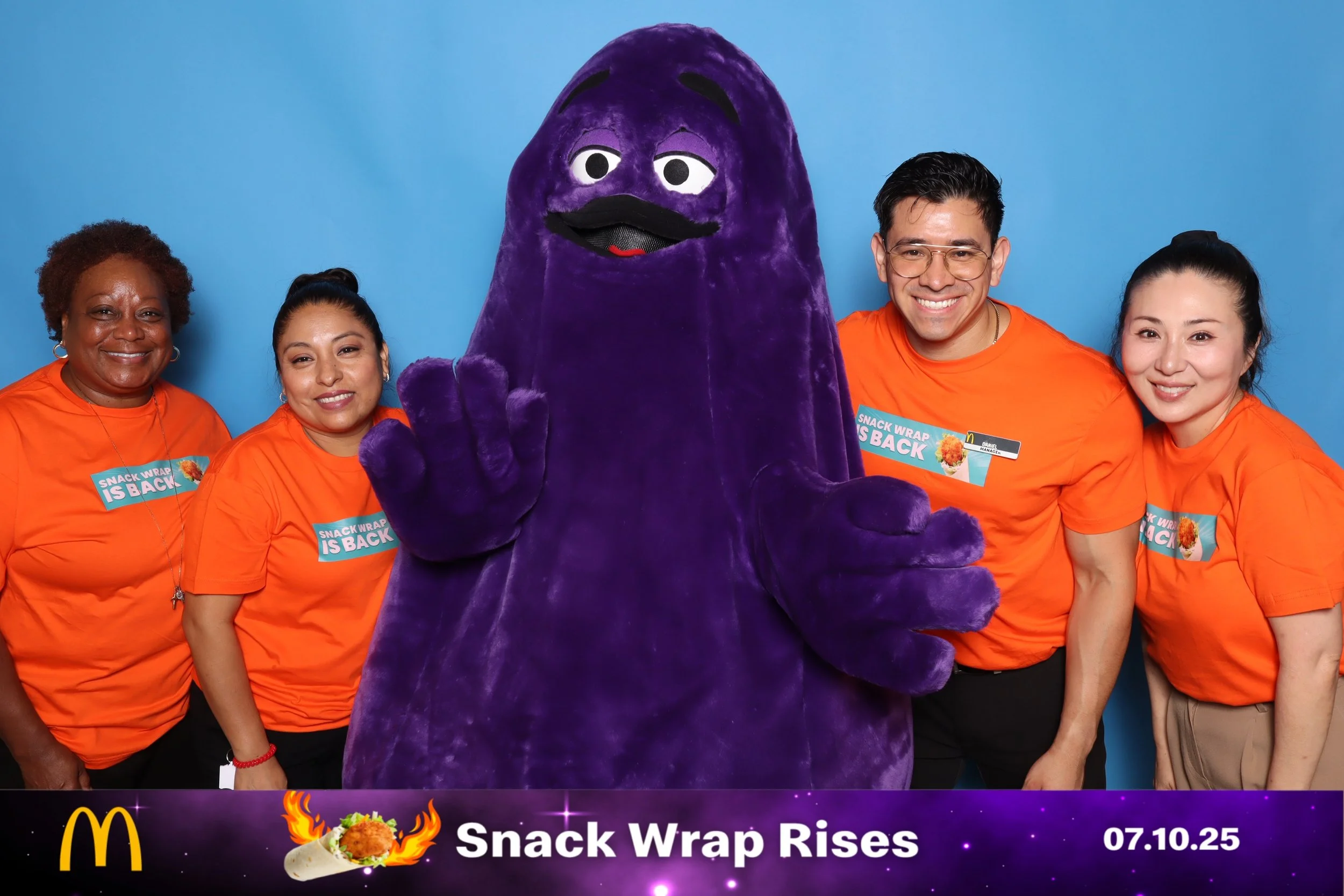 Group of four people in orange shirts standing beside a purple mascot with a mustache and hand raised, with a blue background and a McDonald's logo, celebrating the return of snack wrap with the text 'Snack Wrap Rises' and the date 07.10.25 on a banner at the bottom.