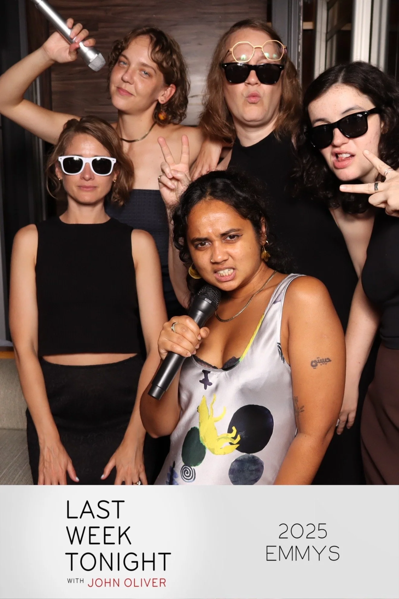 A group of six women at a party or event, some wearing sunglasses and making peace signs, with one woman in front holding a microphone, all smiling or making expressive faces, in a cozy indoor setting.