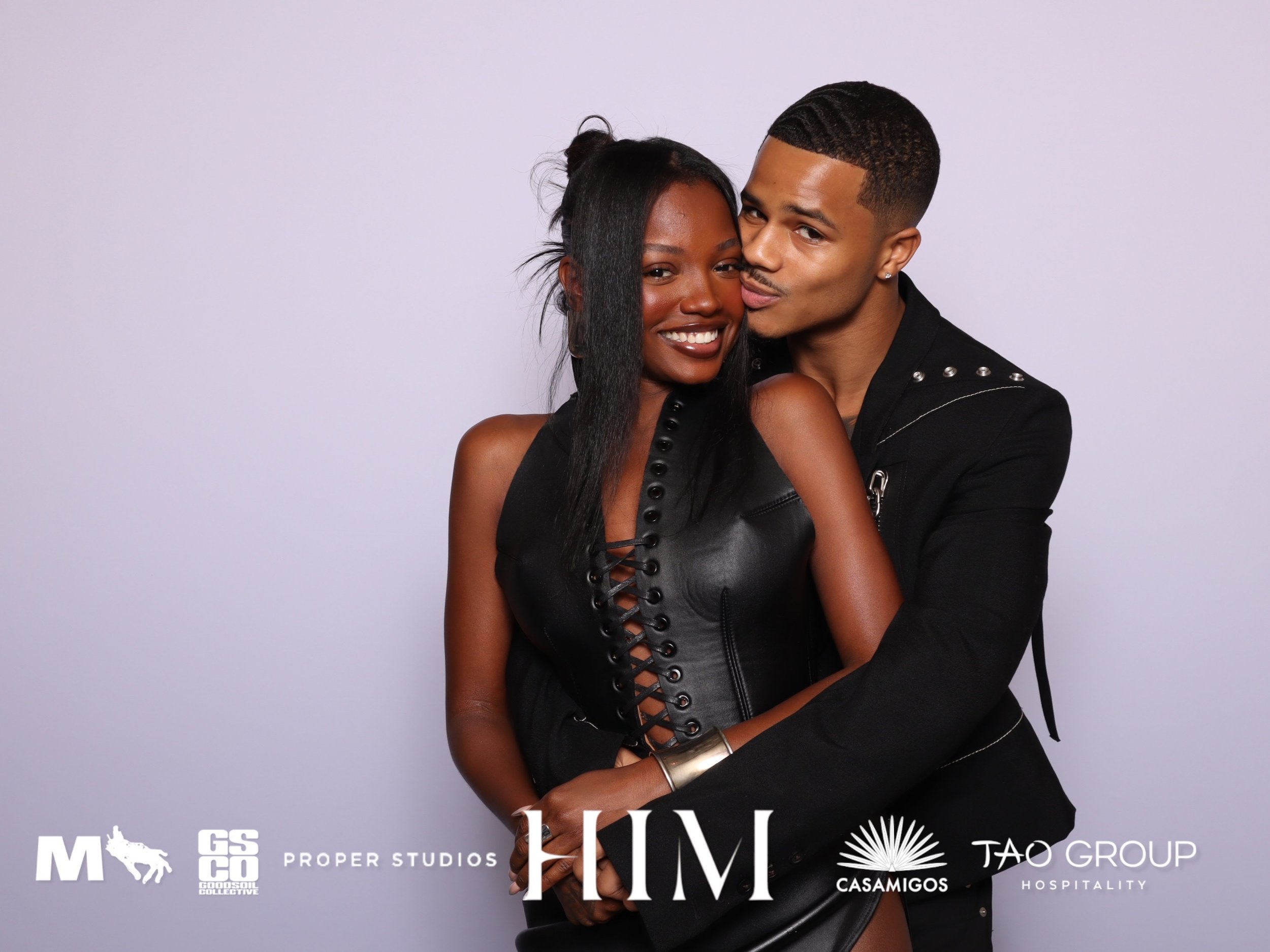 A smiling couple of African American descent, woman and man, embracing each other against a plain light background, wearing black outfits.