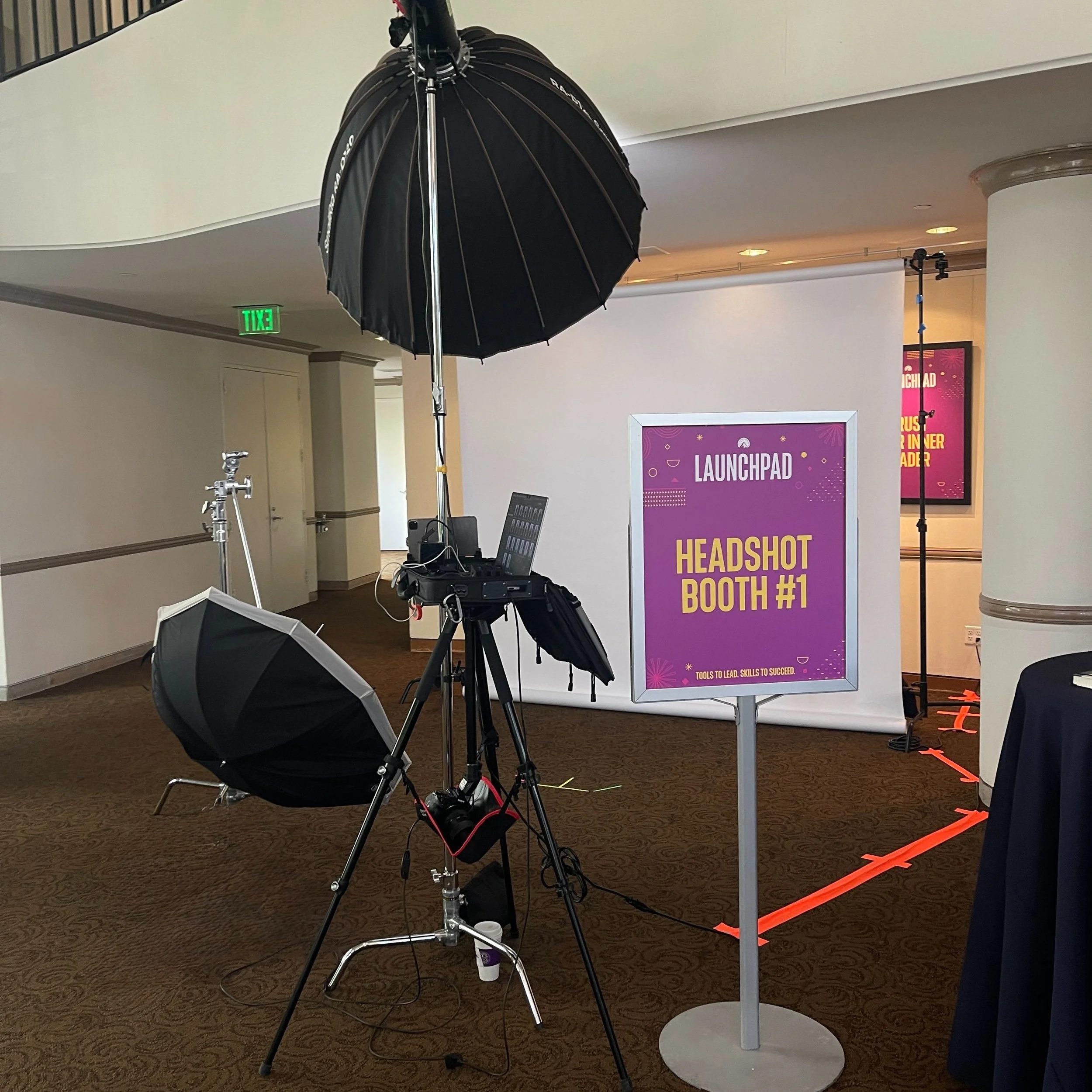 Setup for event photography, including a camera on a tripod with a large black umbrella light and a smaller reflector light, with a sign indicating 'Launchpad Headshot Booth #1' and a purple background.