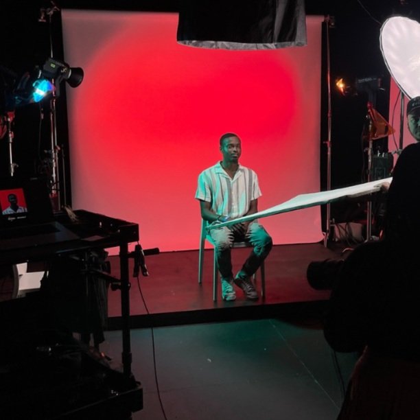 A young man sitting on a chair in a photography studio with a red backdrop and professional lighting equipment, preparing for a photo shoot.