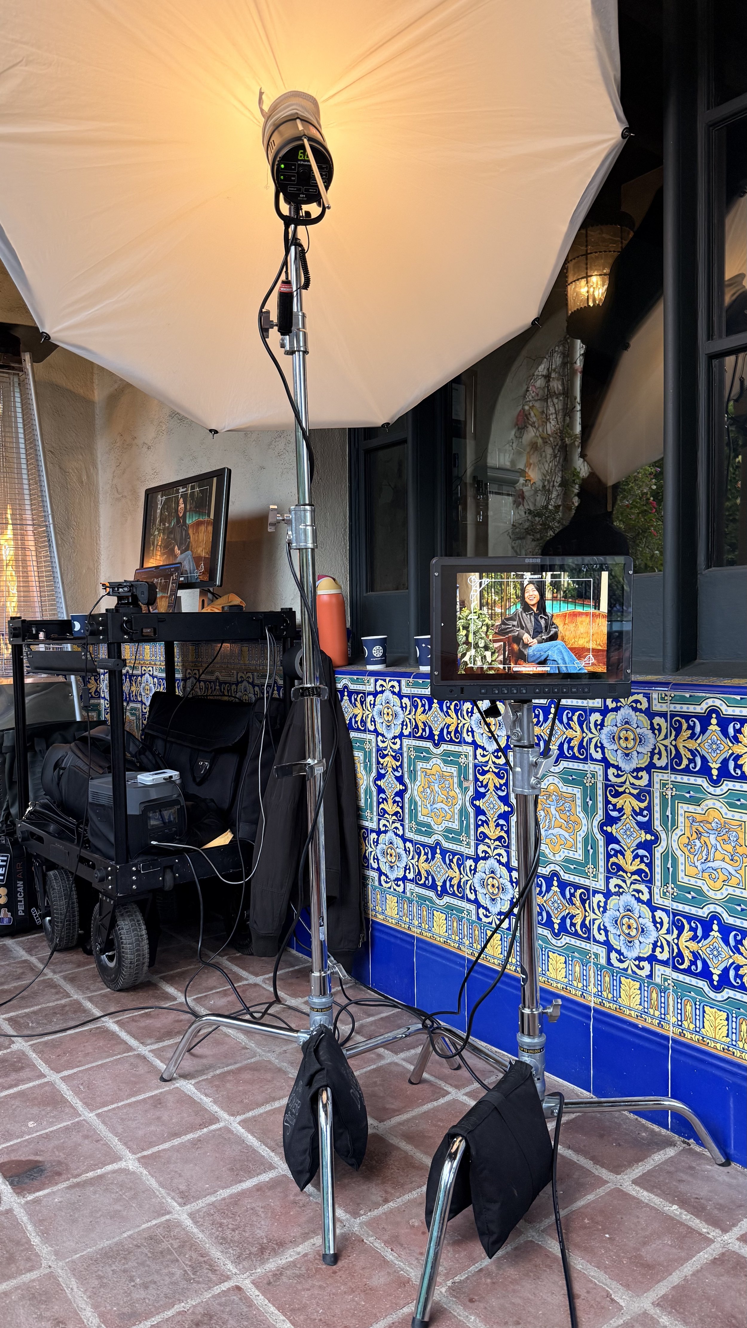 Photo of a photography setup outdoors, including a large umbrella diffuser, a monitor displaying a woman sitting outdoors, and various lighting and camera equipment on a tile patio with decorative blue and yellow tiles.