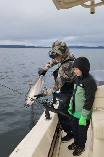 Two people on a boat fishing, one adult and one child, with the adult holding a fish caught in a net over the water.