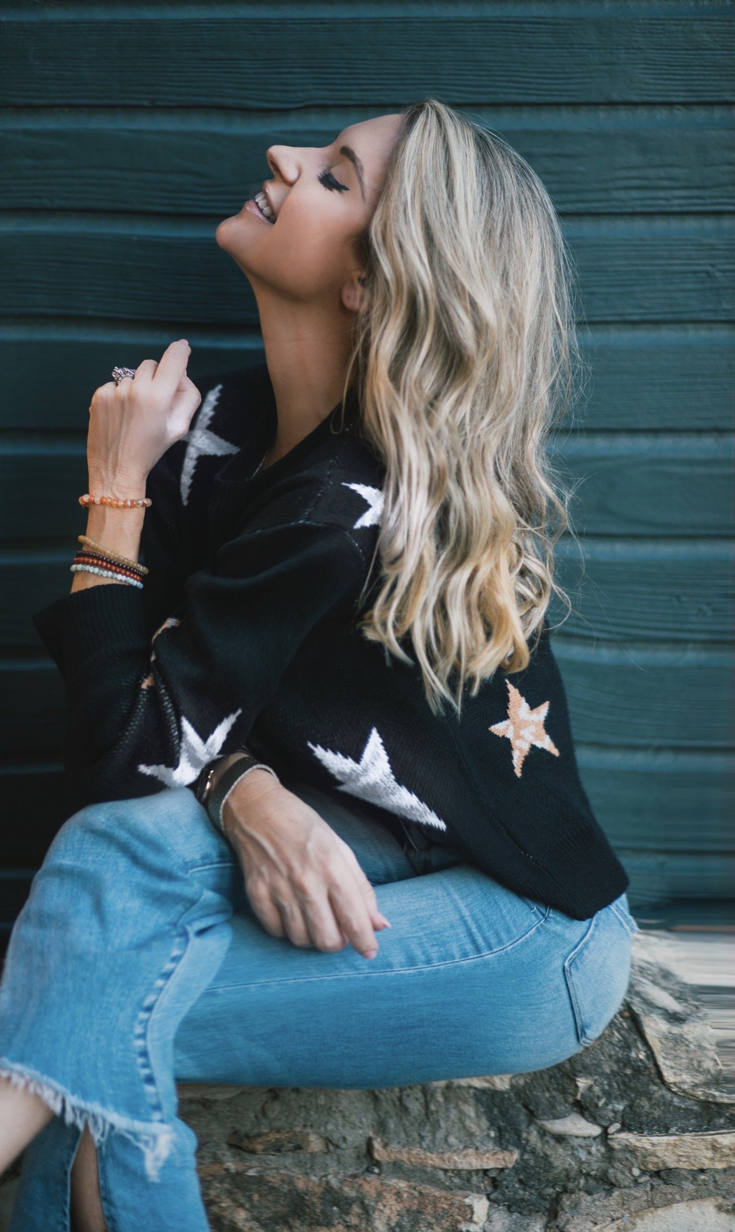 a woman sitting sideways with her head looking up and eyes closed. she teaches how the law of attraction works. she is wearing a navy shirt with white stars. she has blonde hair. the background is a teal colored wood