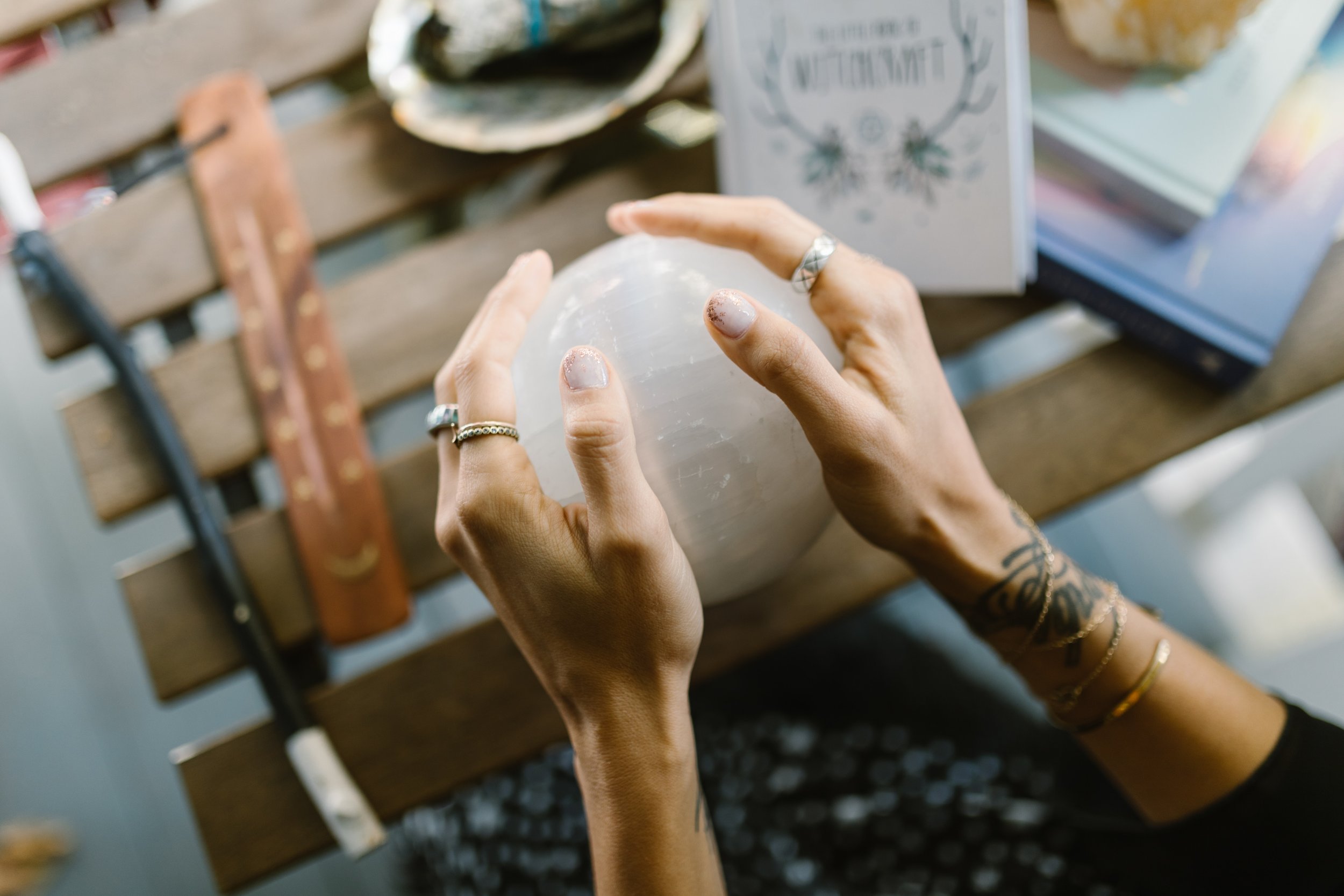 a woman's hands holding onto a crystal ball while she practices mediumship development. The ball is sitting on a wooden table with various spiritual objects like books and incense.
