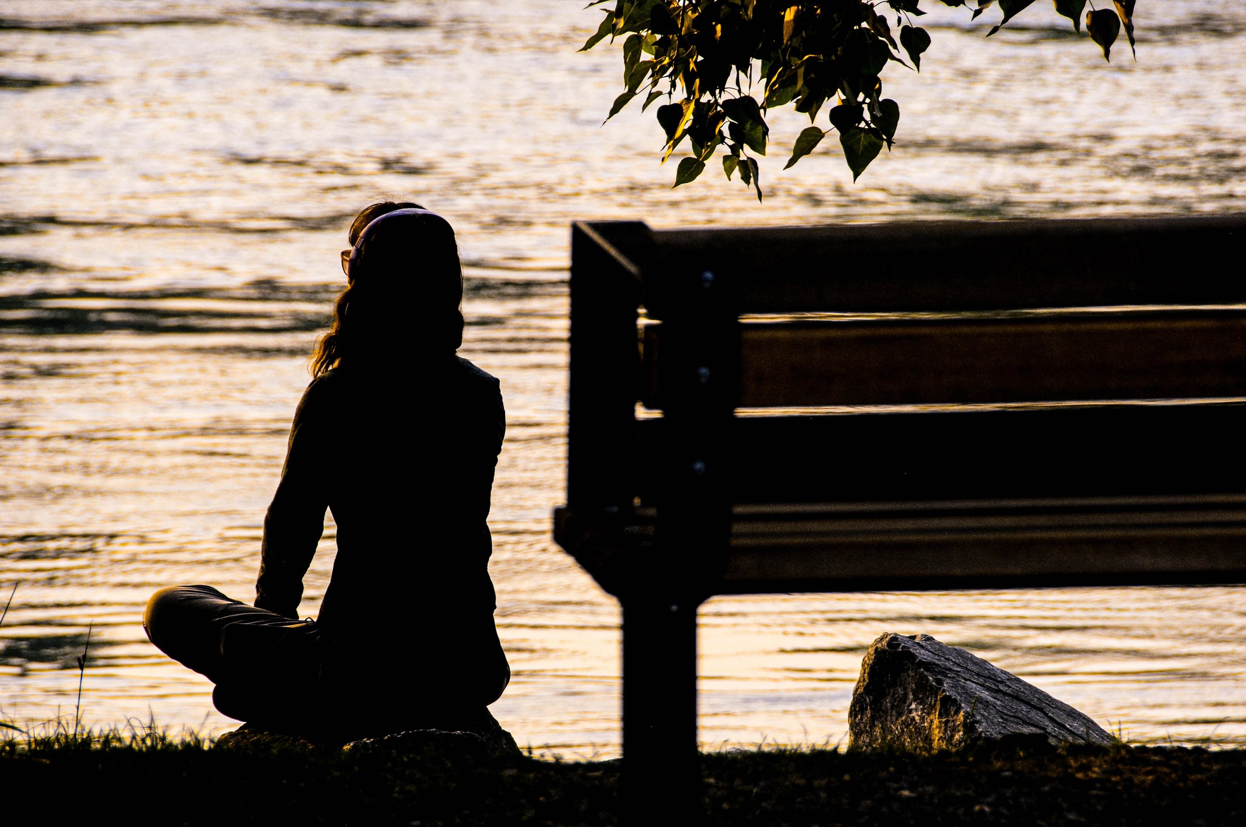 a woman sitting on a rock overlooking a wavy lake at sunset. there is a bench behind and to the right of her, and a leaves from a tree hanging over the bench.