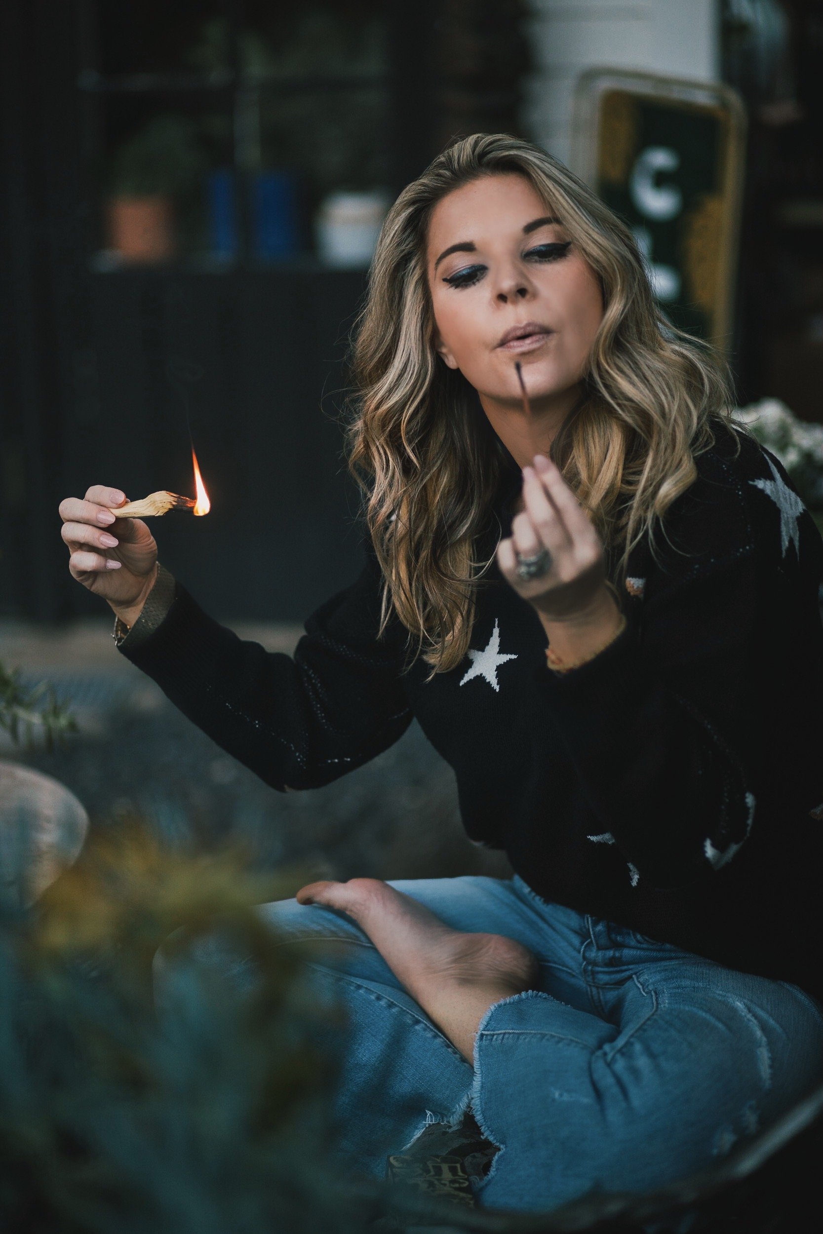 a woman sitting in the grass blowing out a match she used to light some palo santo. she has blonde hair and a navy shirt with white stars. she teaches how the law of attraction works
