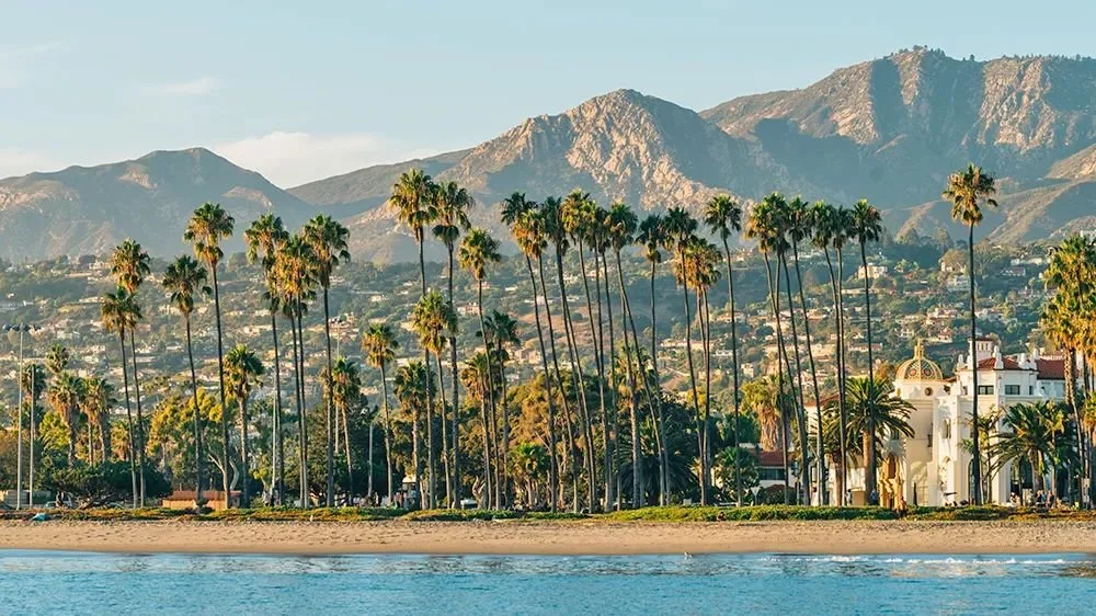 A scenic view of a beach with palm trees, a white building, and mountains in the background.