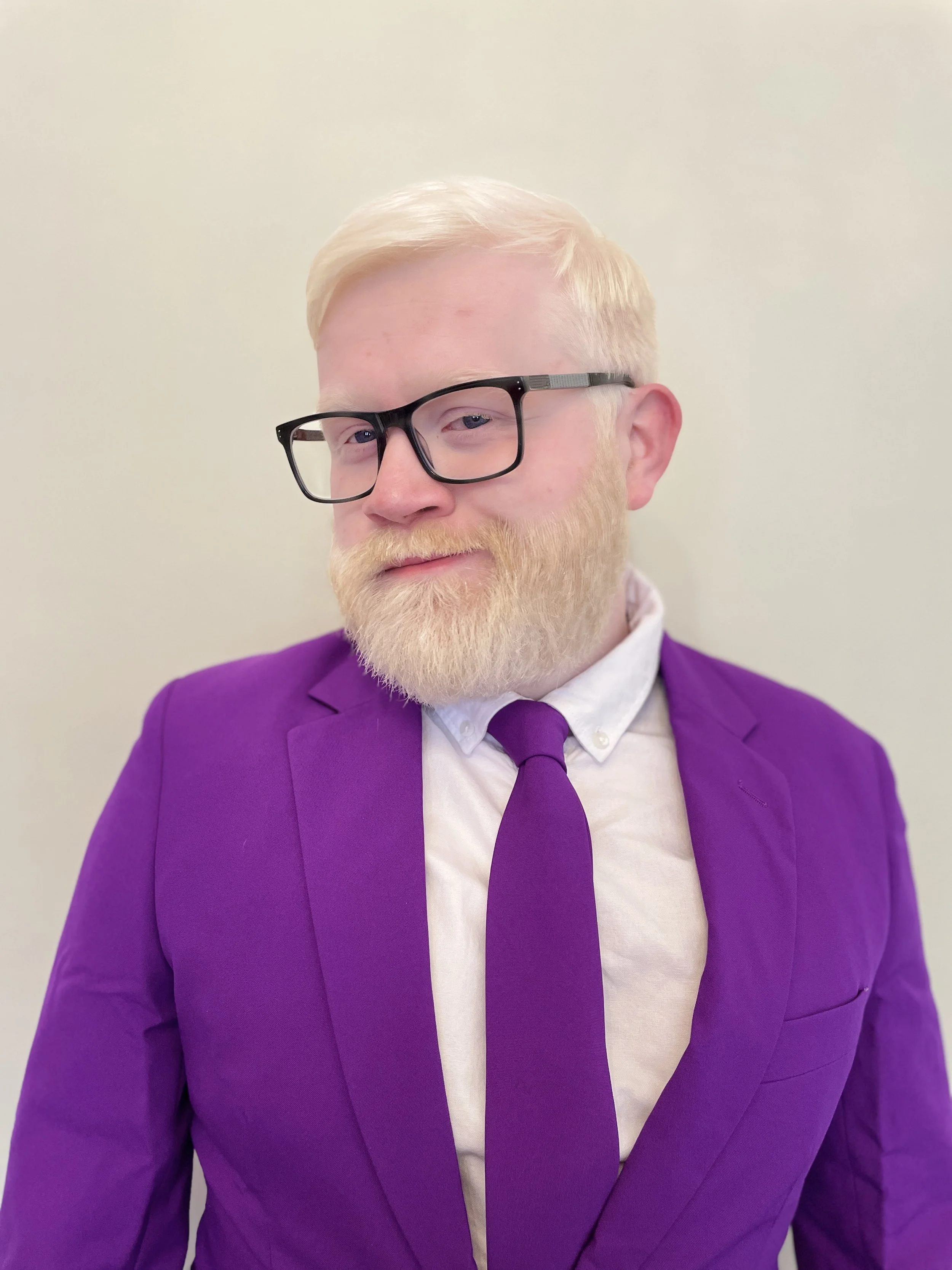 Chris - Has short combed over white hair with thick black framed glasses and white beard. Wearing bright purple suit blazer with matching tie and white shirt underneath.