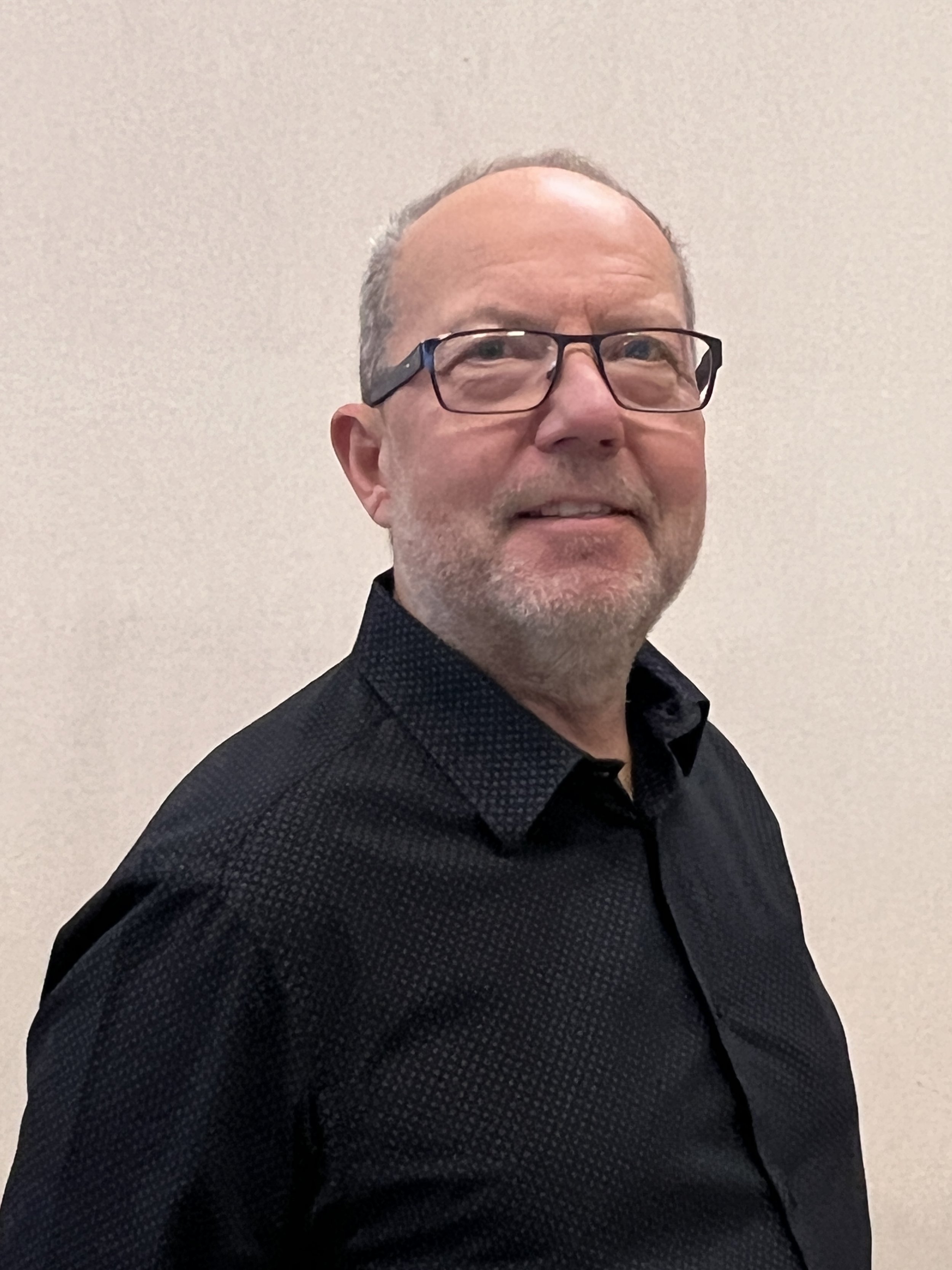 Person with glasses and beard, wearing a dark shirt, standing against a light background.