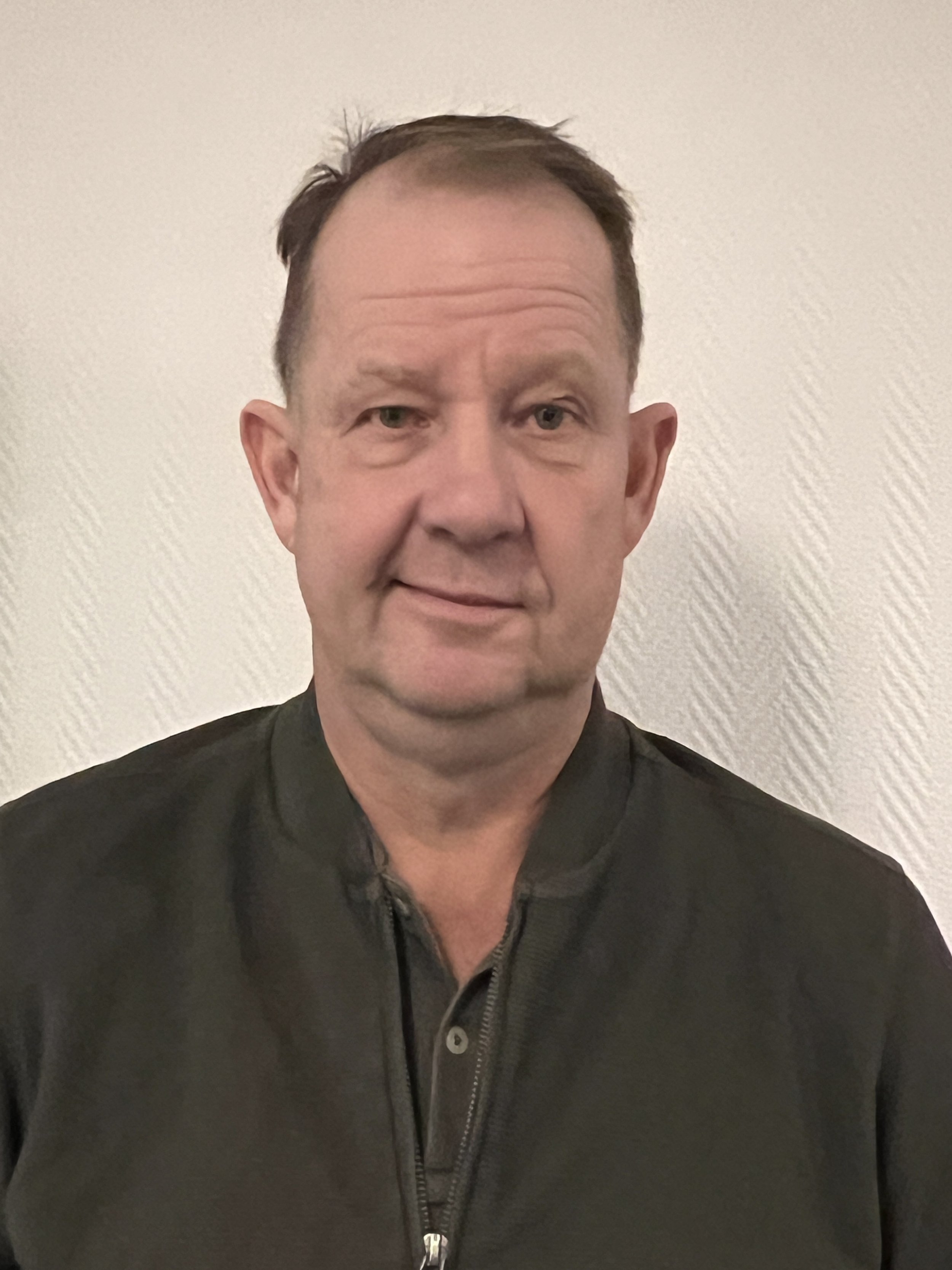 Portrait of a man with short hair and dark sweater, white wall in the background.