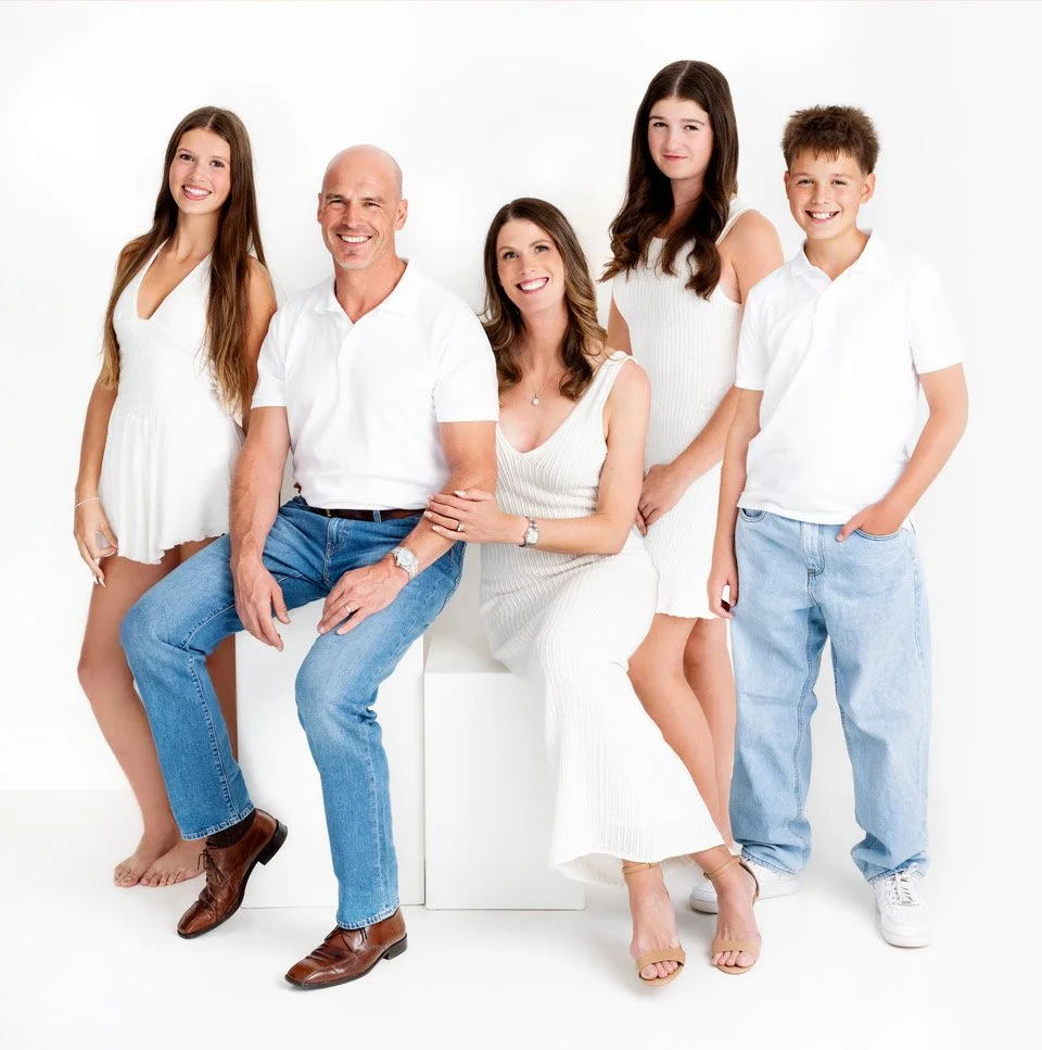 A family of five, including two adult women, one adult man, one teenage girl, and one young boy, dressed in white shirts and light-colored clothing, standing against a white background.