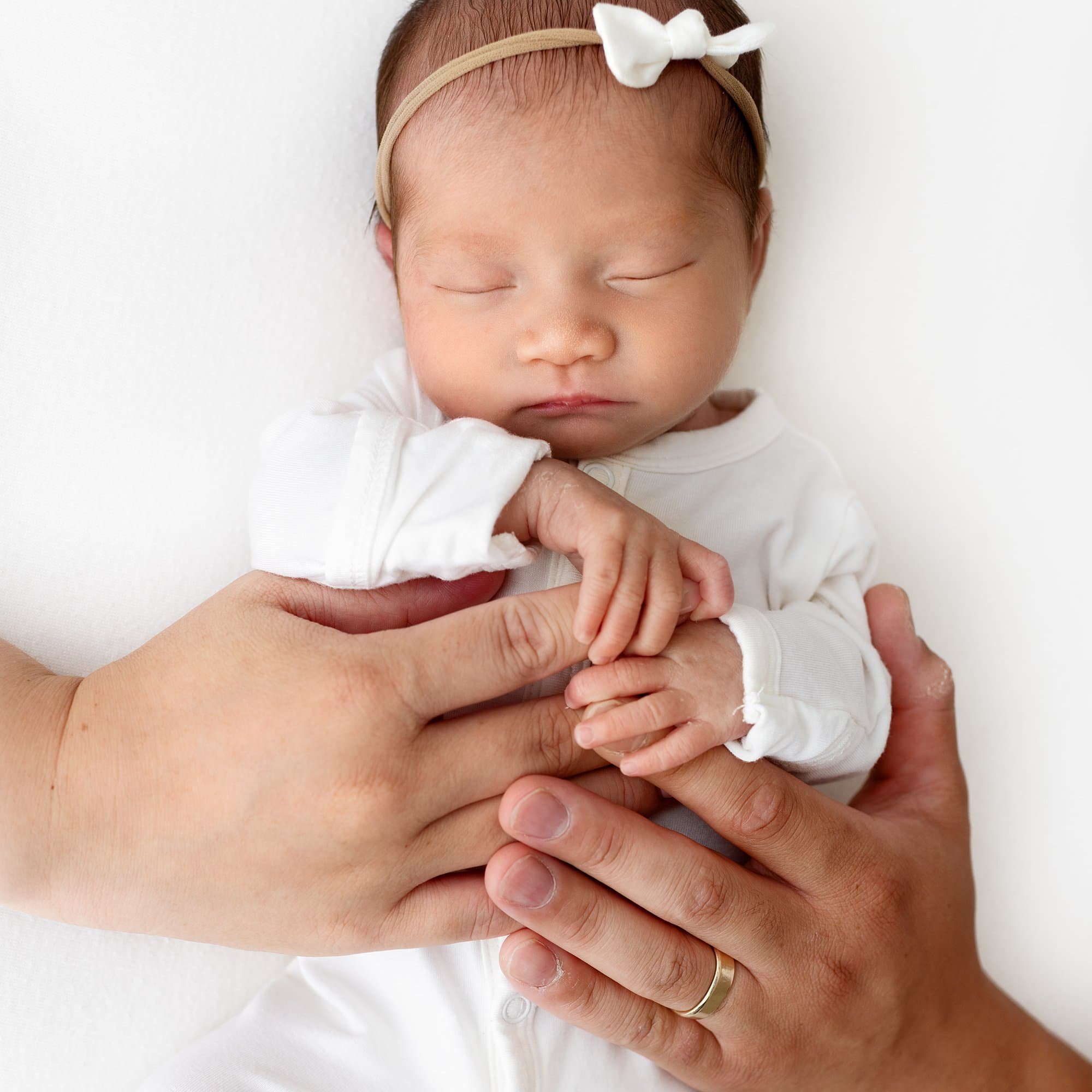 Newborn baby in white outfit and headband, gently holding hands with two adults on a white background.