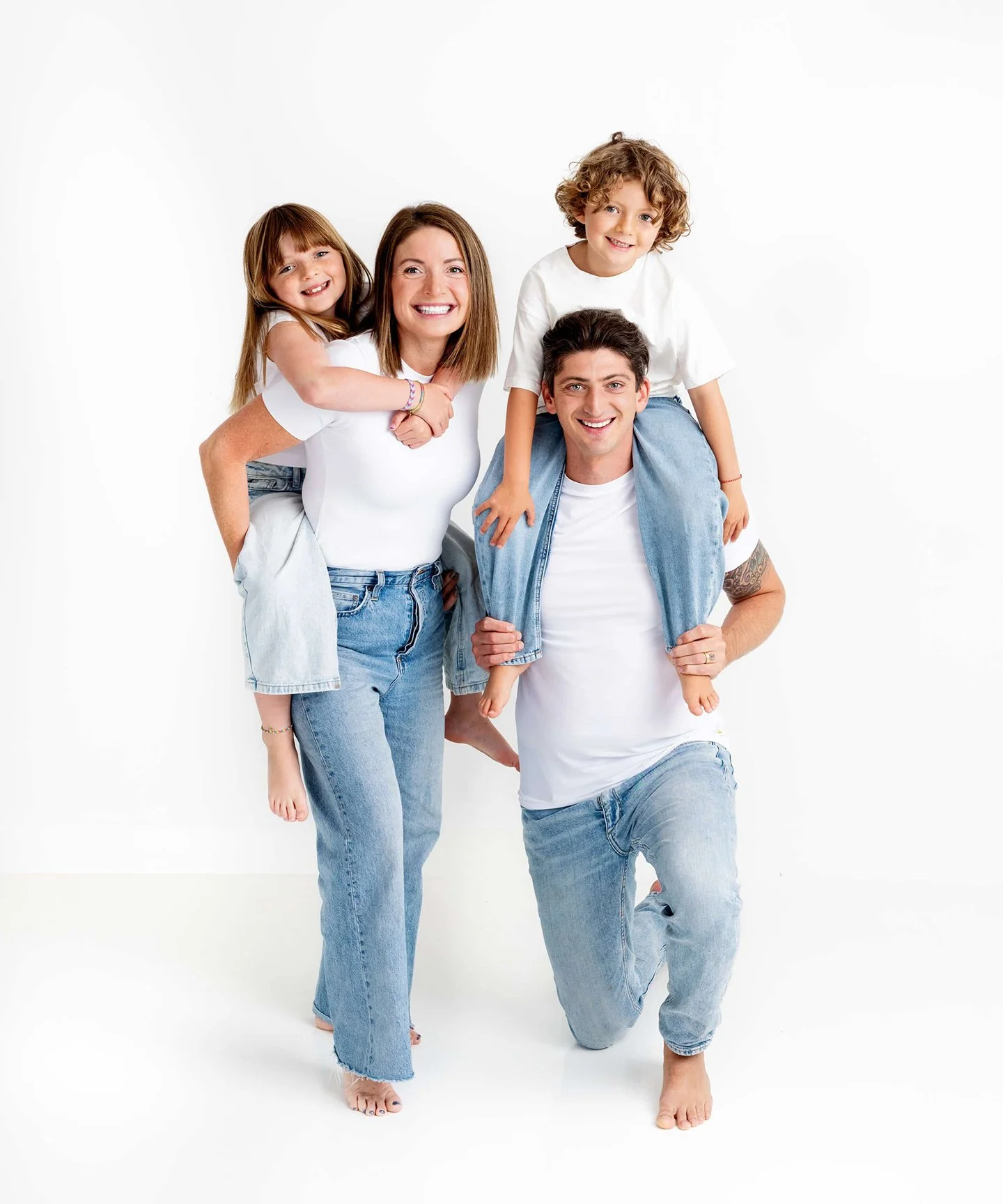 A family of four smiling, with the father kneeling and carrying a young boy, the mother standing and carrying a young girl on her back, all dressed casually in white shirts and jeans against a white background.