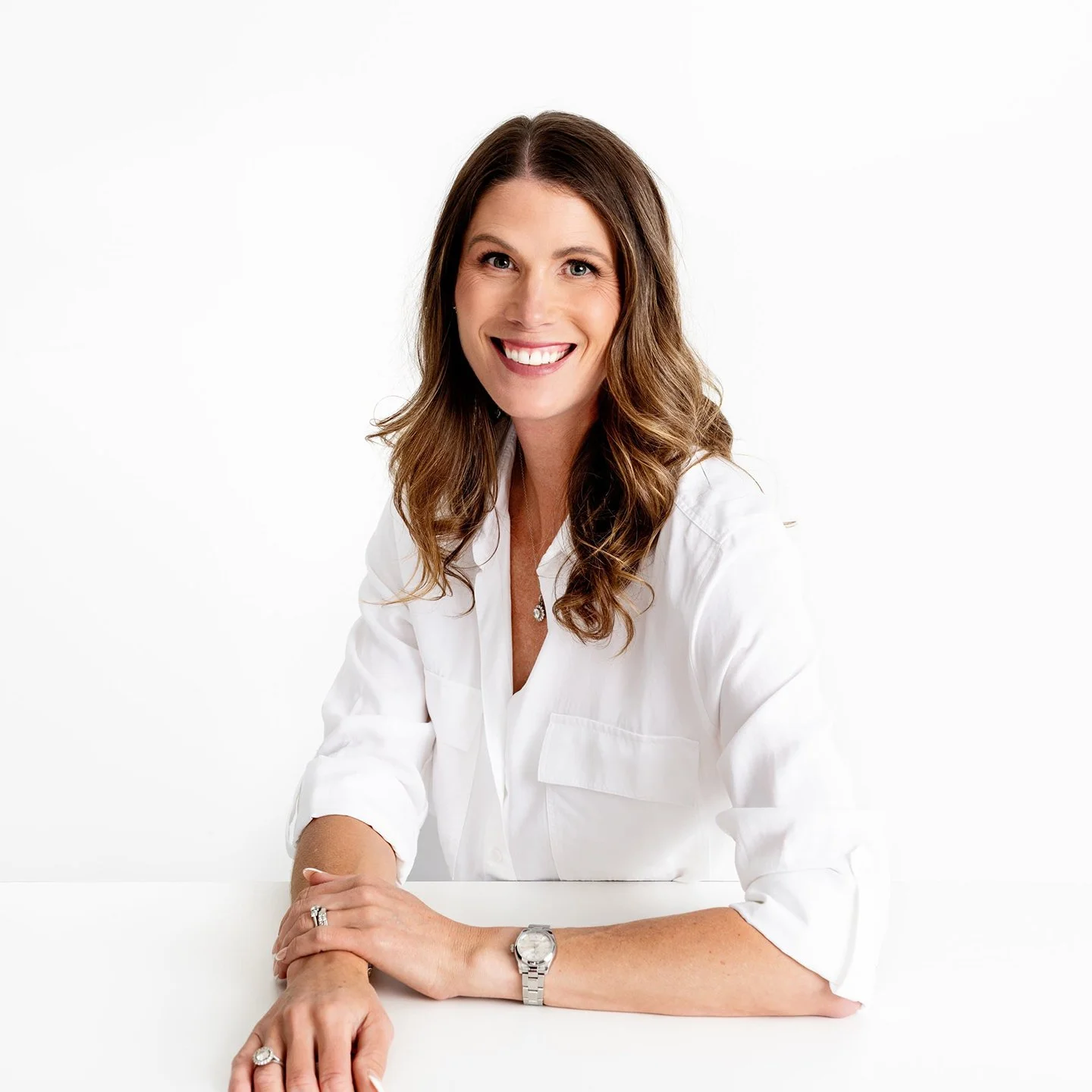 Smiling woman with brown wavy hair wearing a white shirt, sitting at a white table against a white background.