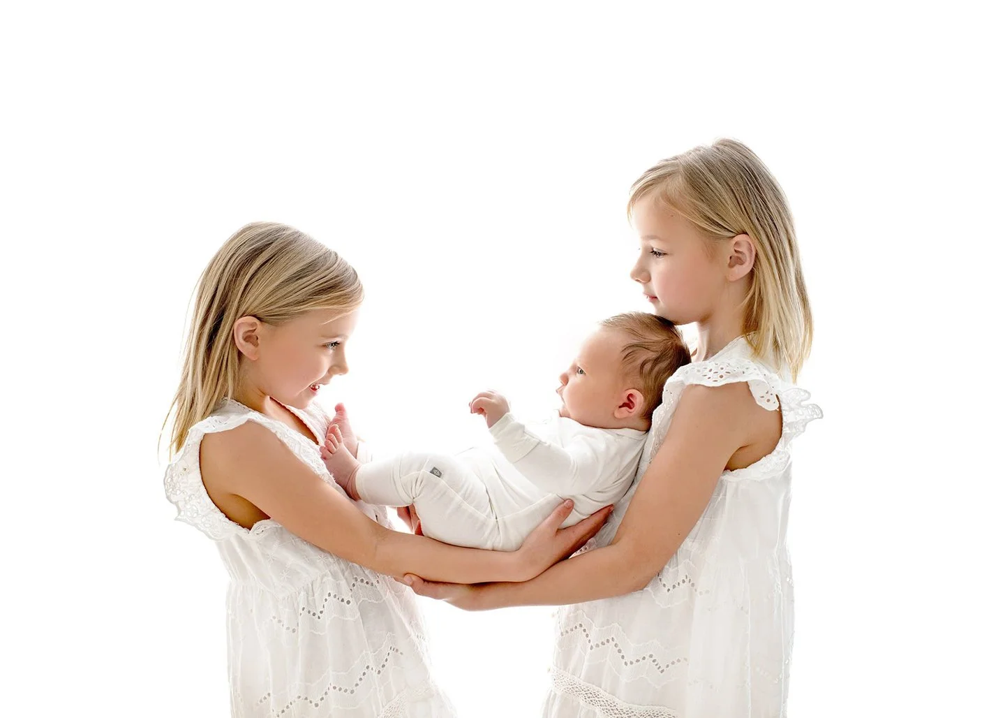 Two young girls in white dresses hold a newborn, capturing a tender moment of baby and family photography against a white background.