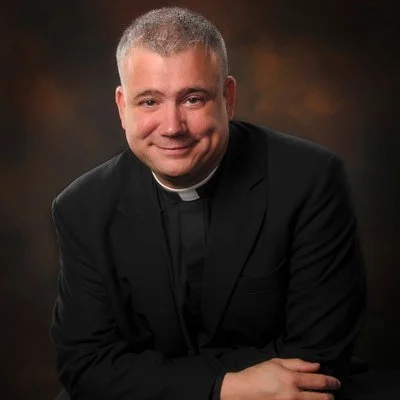 Portrait of a young man in clergy attire with short gray hair, smiling against a dark, blurred background.