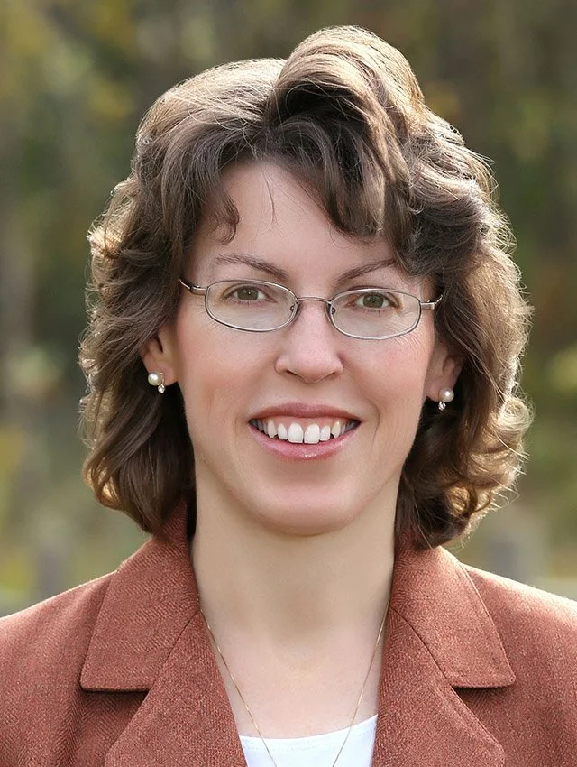 A woman with wavy brown hair and glasses smiling outdoors, wearing pearl earrings and a brown blazer.