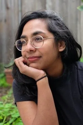 Swamy wearing round glasses and a black t-shirt, leaning with her chin resting on her hand