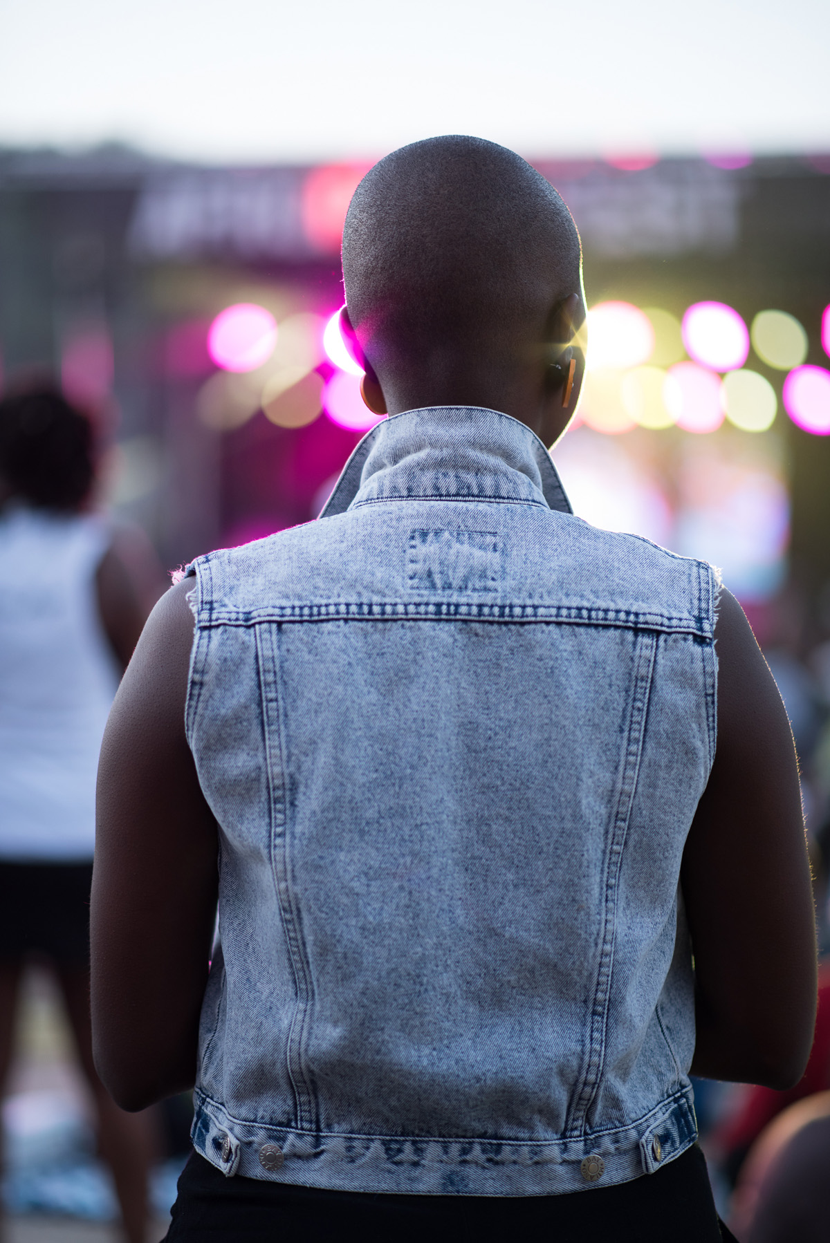 Person wearing a denim vest at an outdoor concert or event, back view.