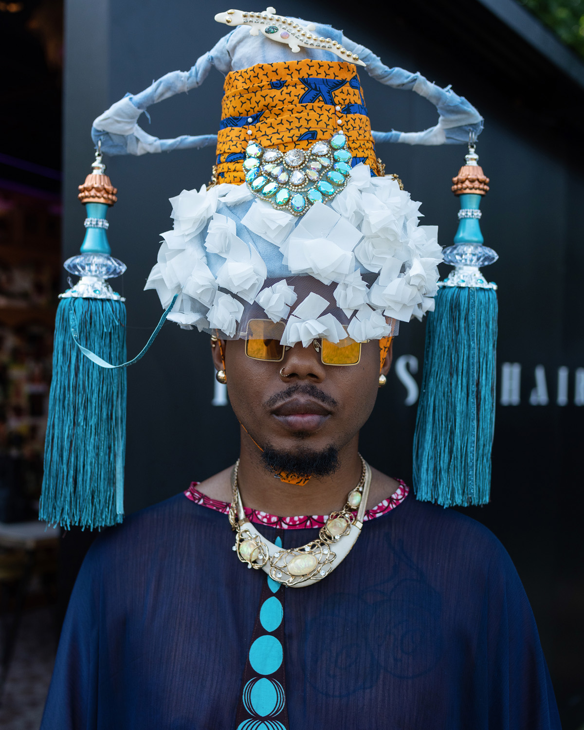 Person wearing an elaborate headdress with blue tassels, white and orange designs, sunglasses, and jewelry.