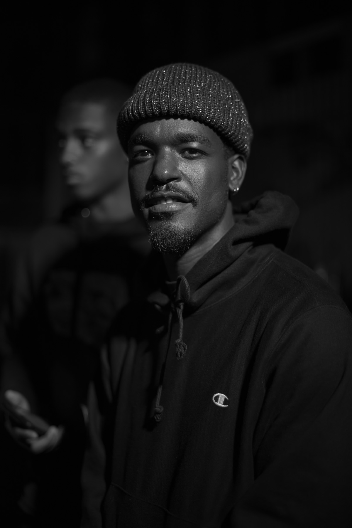 Black and white portrait of a man wearing a beanie and a hooded sweatshirt, standing outdoors at night.
