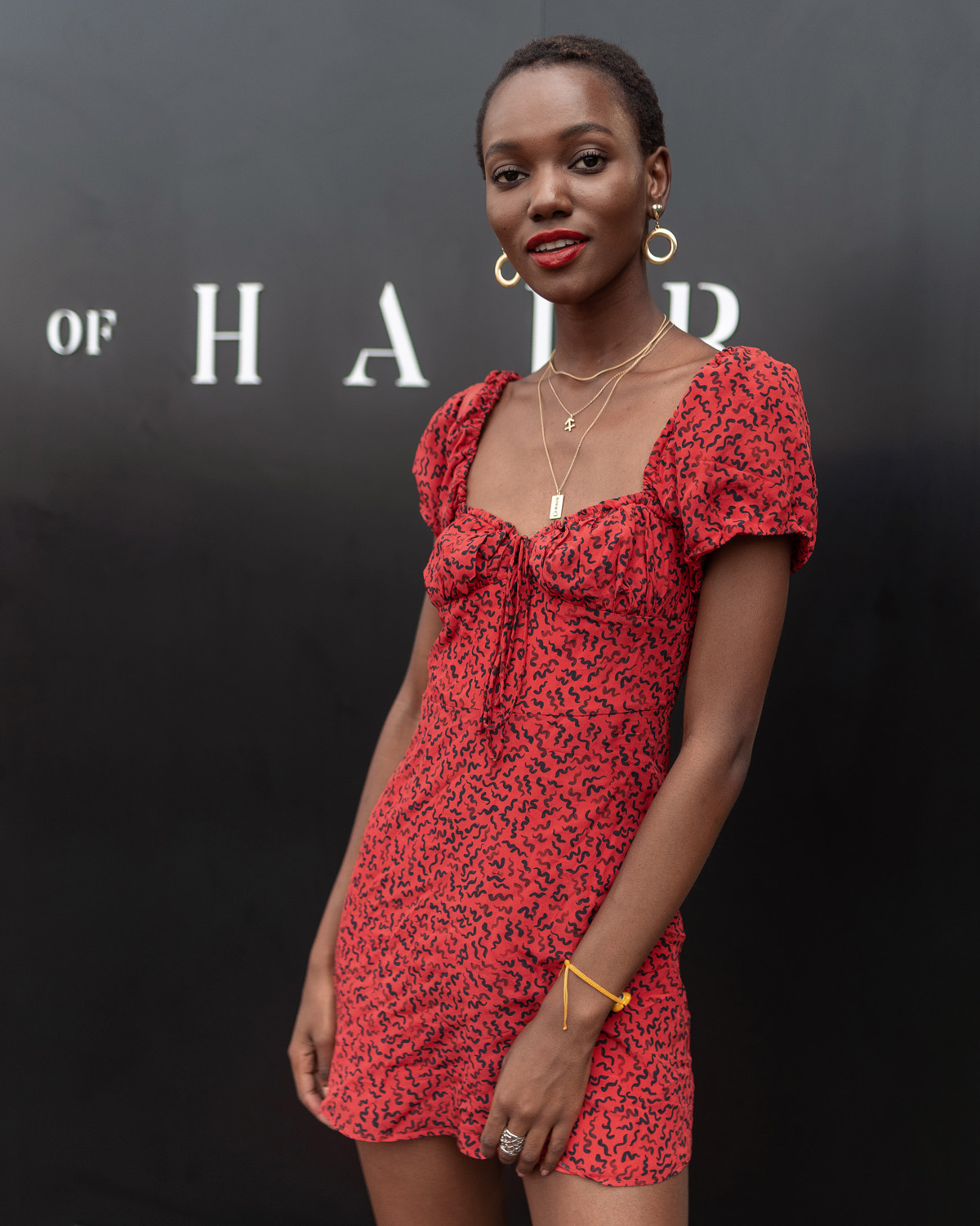 Woman in a red patterned dress with necklaces and hoop earrings, standing against a dark background with partially visible text. She is smiling slightly and has arms by her sides.