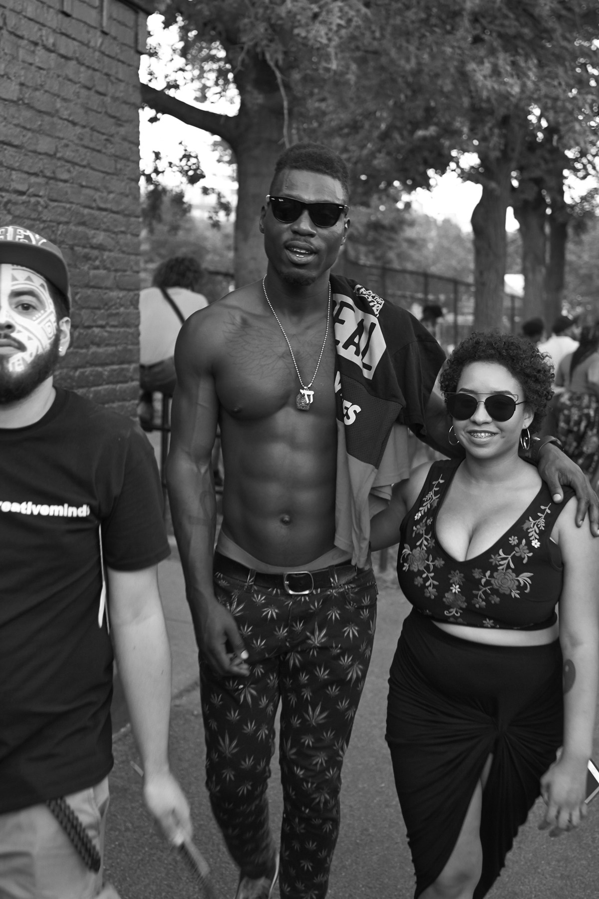 Three people walking outdoors, one man shirtless wearing sunglasses, holding a towel, another man with face paint, and a woman wearing a patterned top and skirt, all in a park setting.