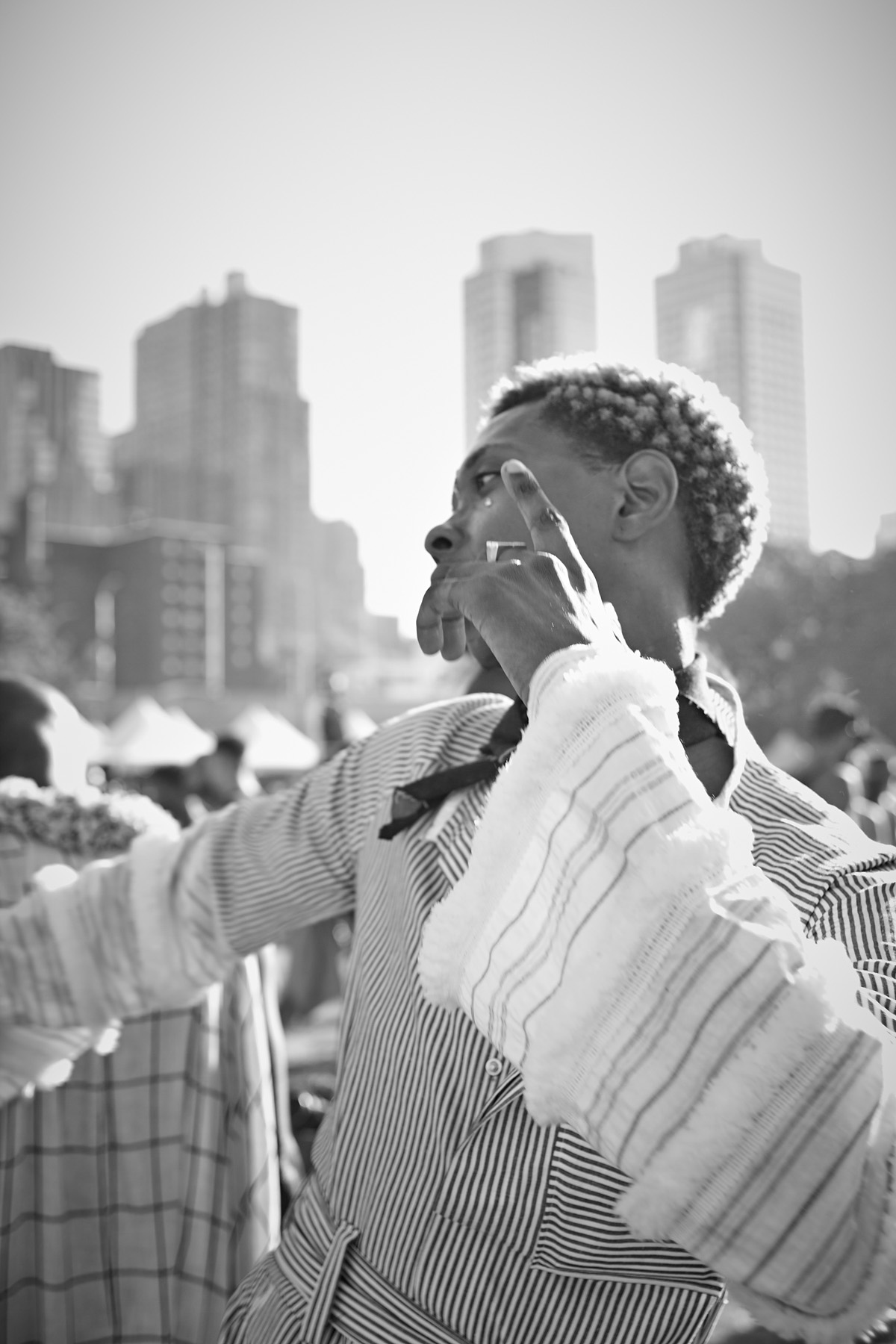 Black and white photo of a person with short curly hair, wearing a striped shirt with flared sleeves, posing with one hand up in an urban setting with tall buildings in the background.