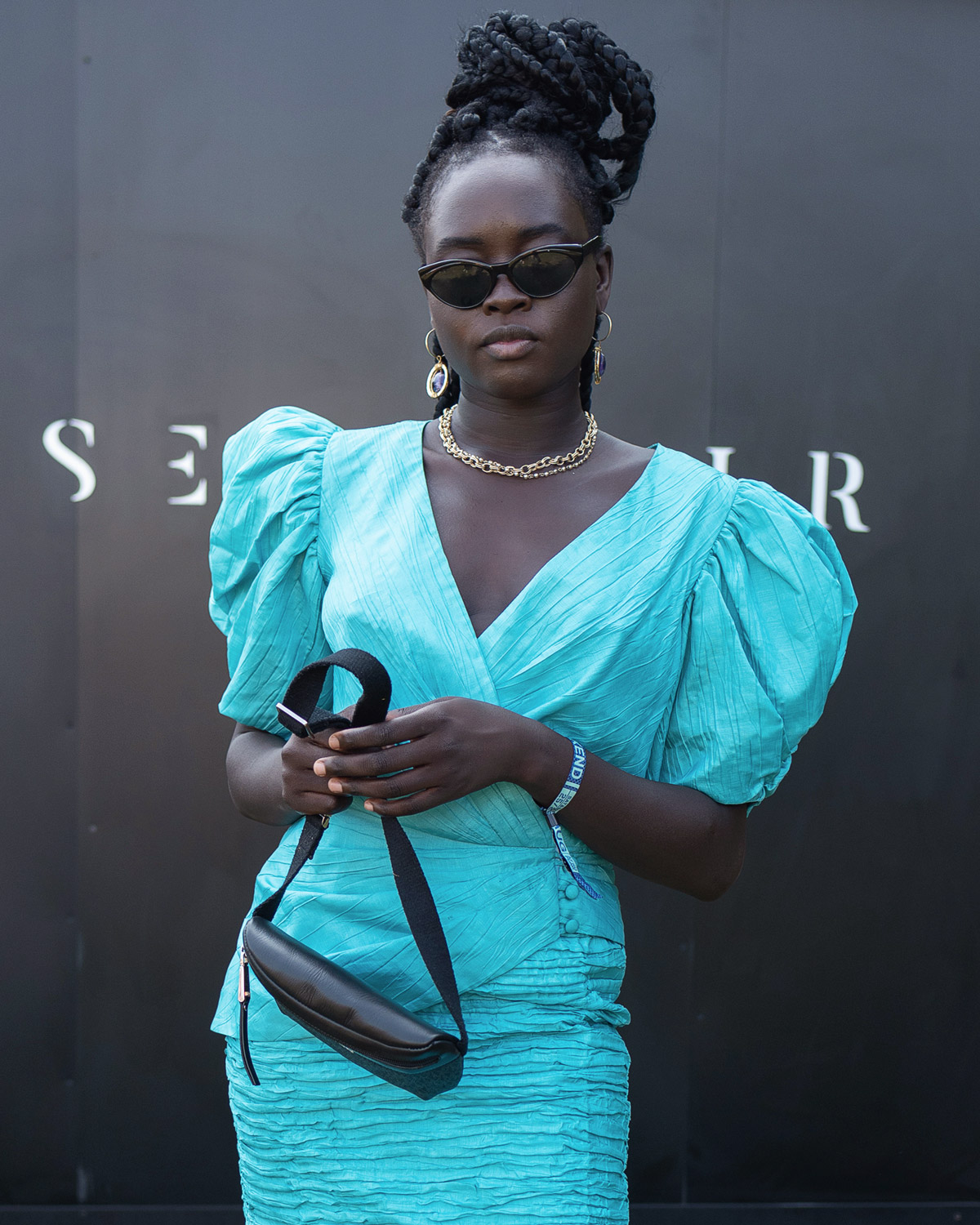 Person in turquoise puff-sleeve dress, holding a black handbag, wearing sunglasses and jewelry, standing in front of a dark background.