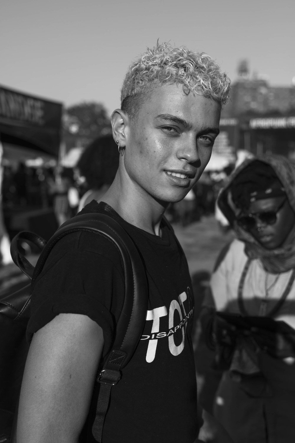 Black and white portrait of a person with curly hair, wearing a t-shirt and backpack, standing outdoors at an event.
