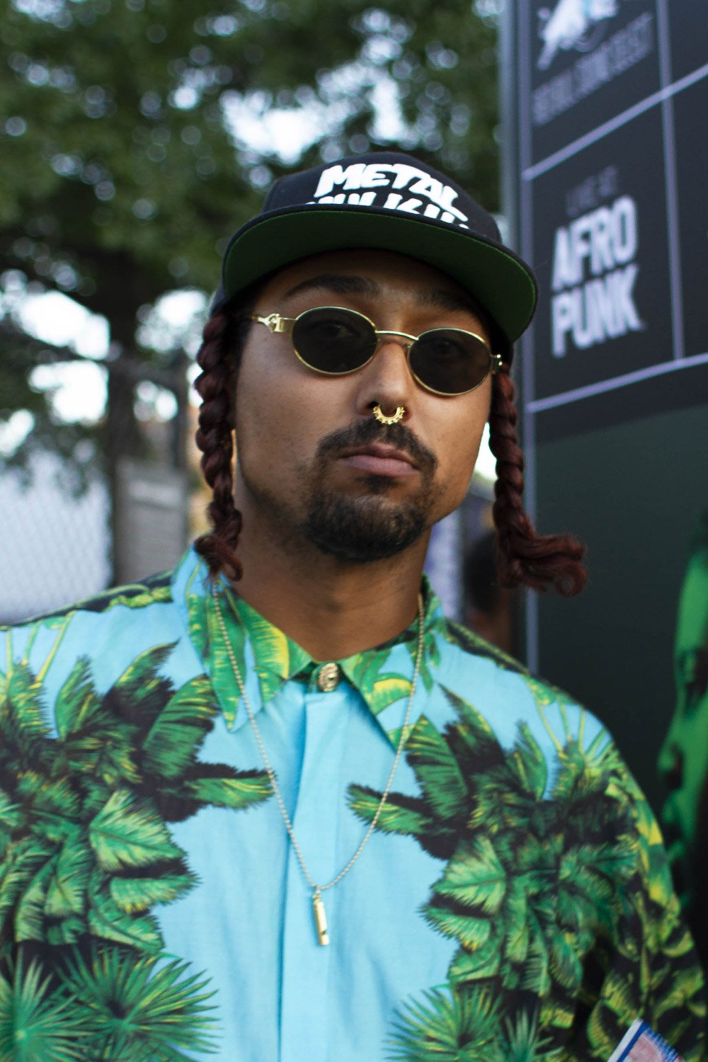 Person wearing sunglasses, cap, nose ring, and floral shirt at a festival.