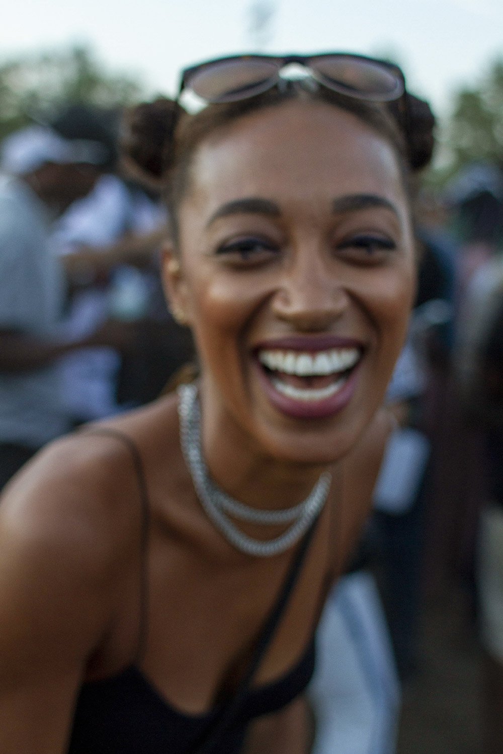 Smiling person with sunglasses on head, wearing a necklace and black top, outdoors.