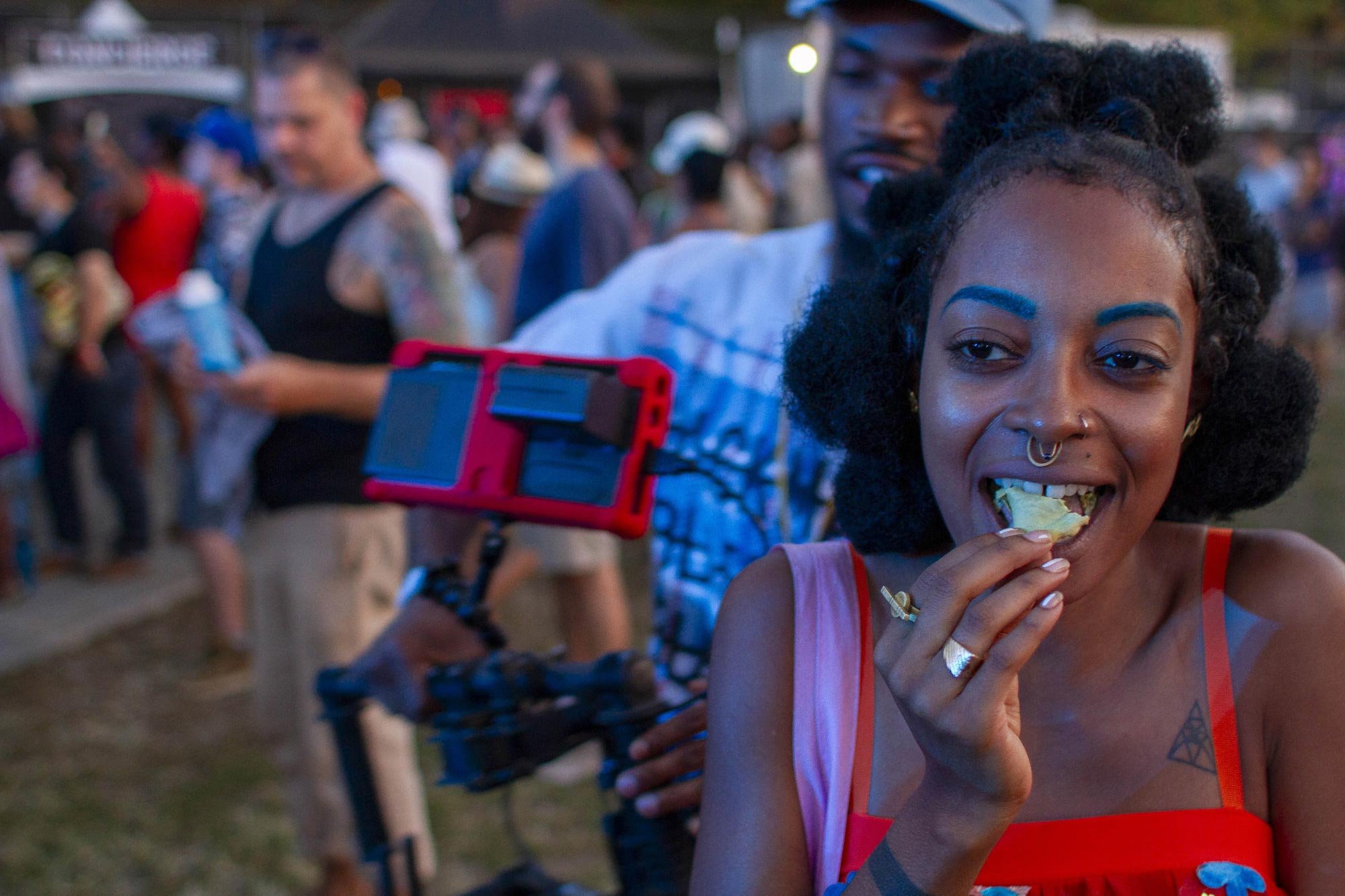 Woman eating at outdoor festival, holding food in hand, surrounded by crowd, with blue eyebrows and nose ring, brightly dressed.