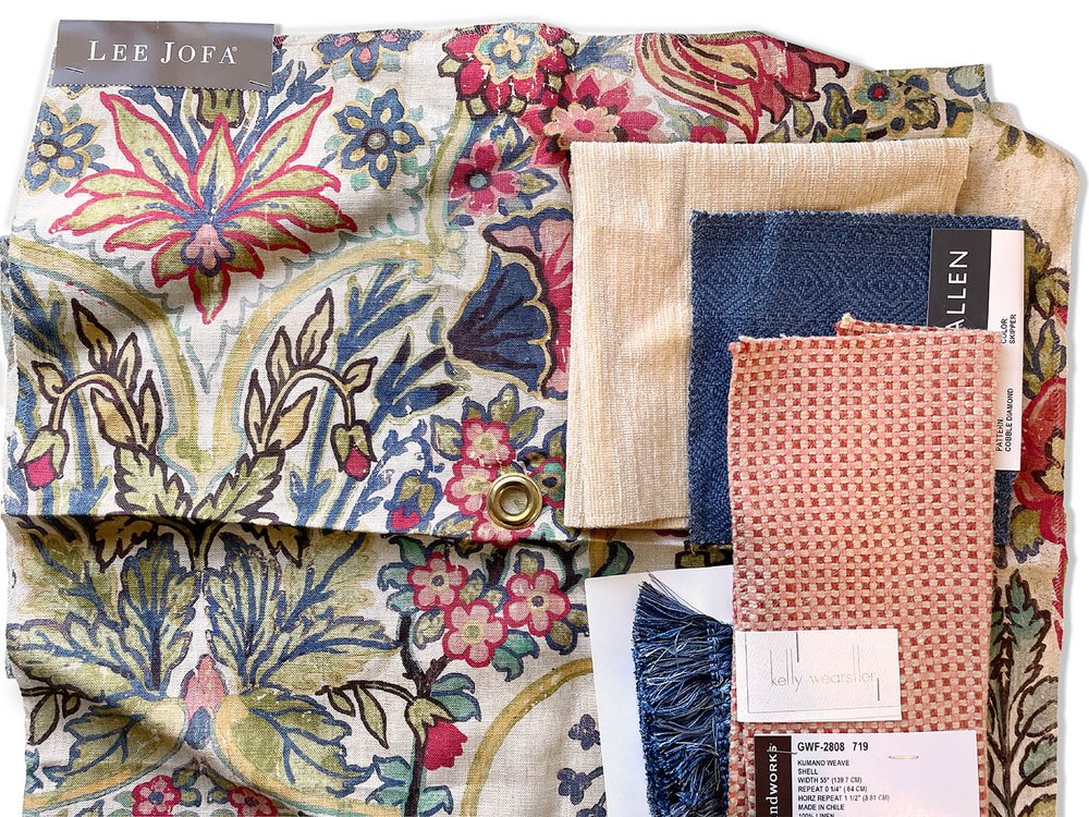 Interior design materials are layered on top of one another. Lee Jofa printed fabric with blue, yellow, red, and green botanicals. Solid off white chenille, solid blue twill, pink and red check, and blue fringe trim.