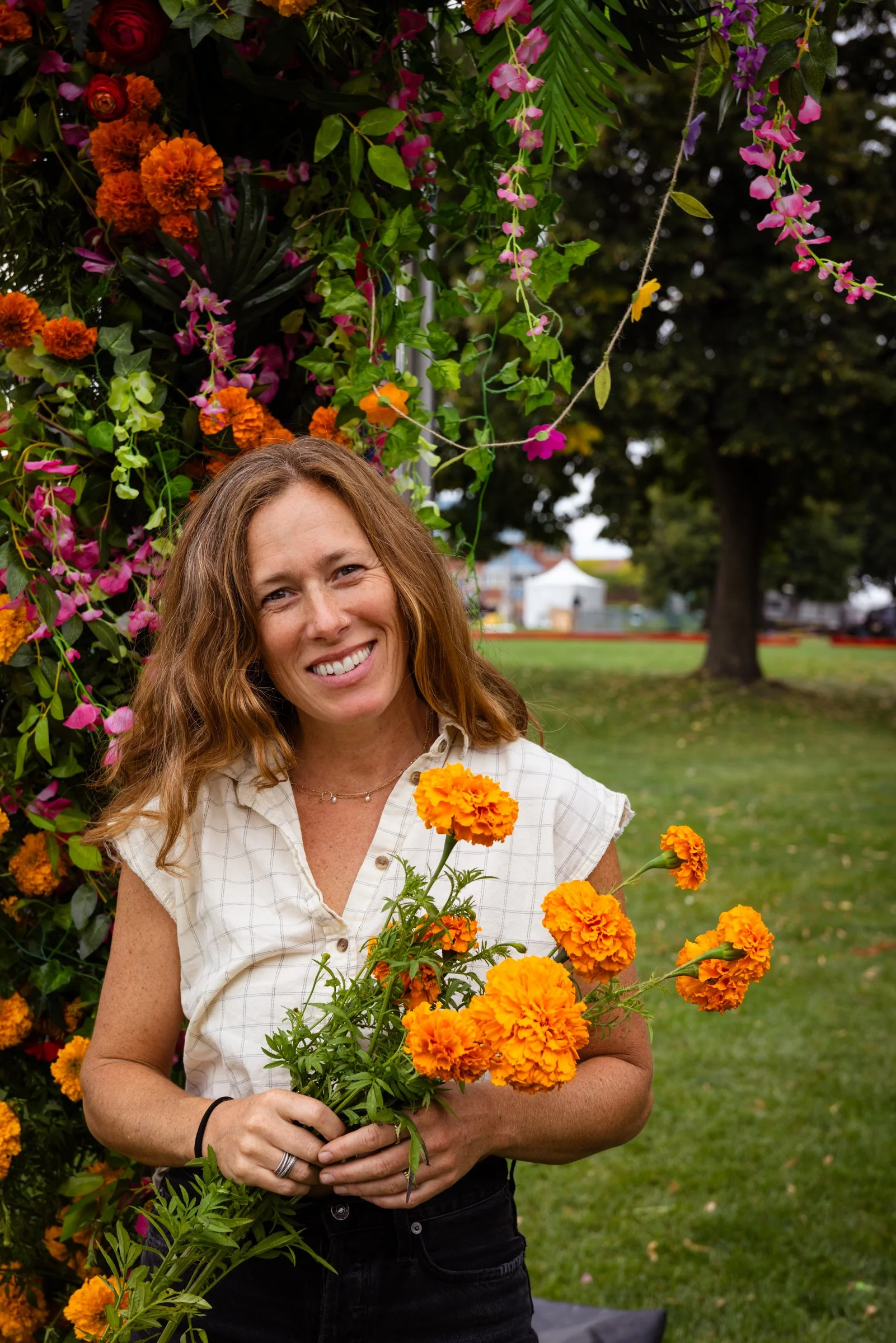 A woman with brown hair, smiling, holding a bouquet of orange marigold flowers. She stands outdoors near a floral arrangement with various flowers and green leaves, with a grassy area and trees in the background.