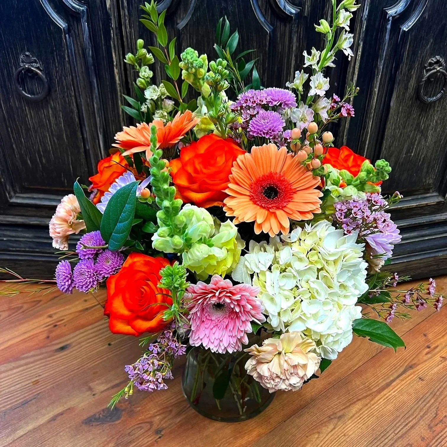 Designed with yellow snapdragon, peach and pink gerbera daisies, orange roses, white hydrangea, peach carnations, purple wax flower, peach hypericum berries and lush greens.